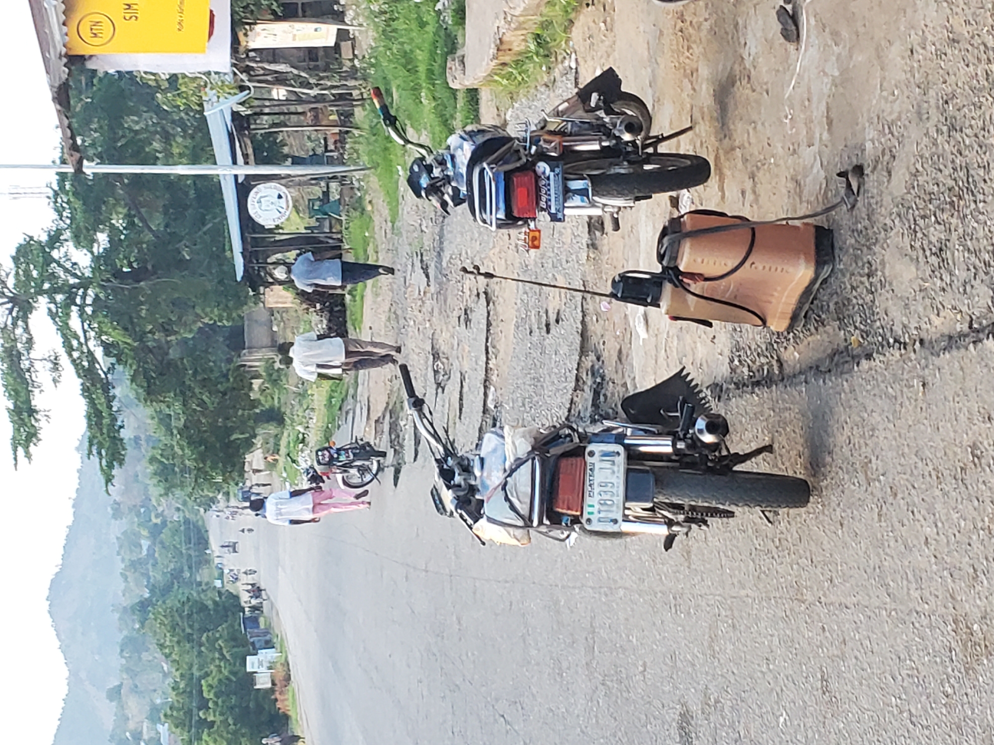 The image shows a street scene in Ampan, Nigeria, with several motorcycles parked along the side of a paved road. Two men, dressed in light-colored shirts and trousers, are standing on the roadside. One man is wearing a white shirt and a dark blue or black trouser, while the other is in a light blue shirt and dark trousers.
In the foreground, two motorcycles are prominently displayed. The one on the right is black with a visible license plate that reads "1839 JJ". Attached to the back of this motorcycle is a tan-colored container with black straps, possibly a fuel tank or sprayer. The second motorcycle, to the left, is also dark-colored and appears to have some items strapped to its rear.
Further down the road, more motorcycles and vehicles are visible, suggesting a busy or populated area. The surrounding environment includes green trees and vegetation on the left side of the image, with a hillside in the background. On the right side of the road, there is a rough, dirt or gravel surface. A yellow sign with black lettering, including the "MTN" logo and the word "SIM," is visible in the upper left corner. The lighting suggests it is daytime.