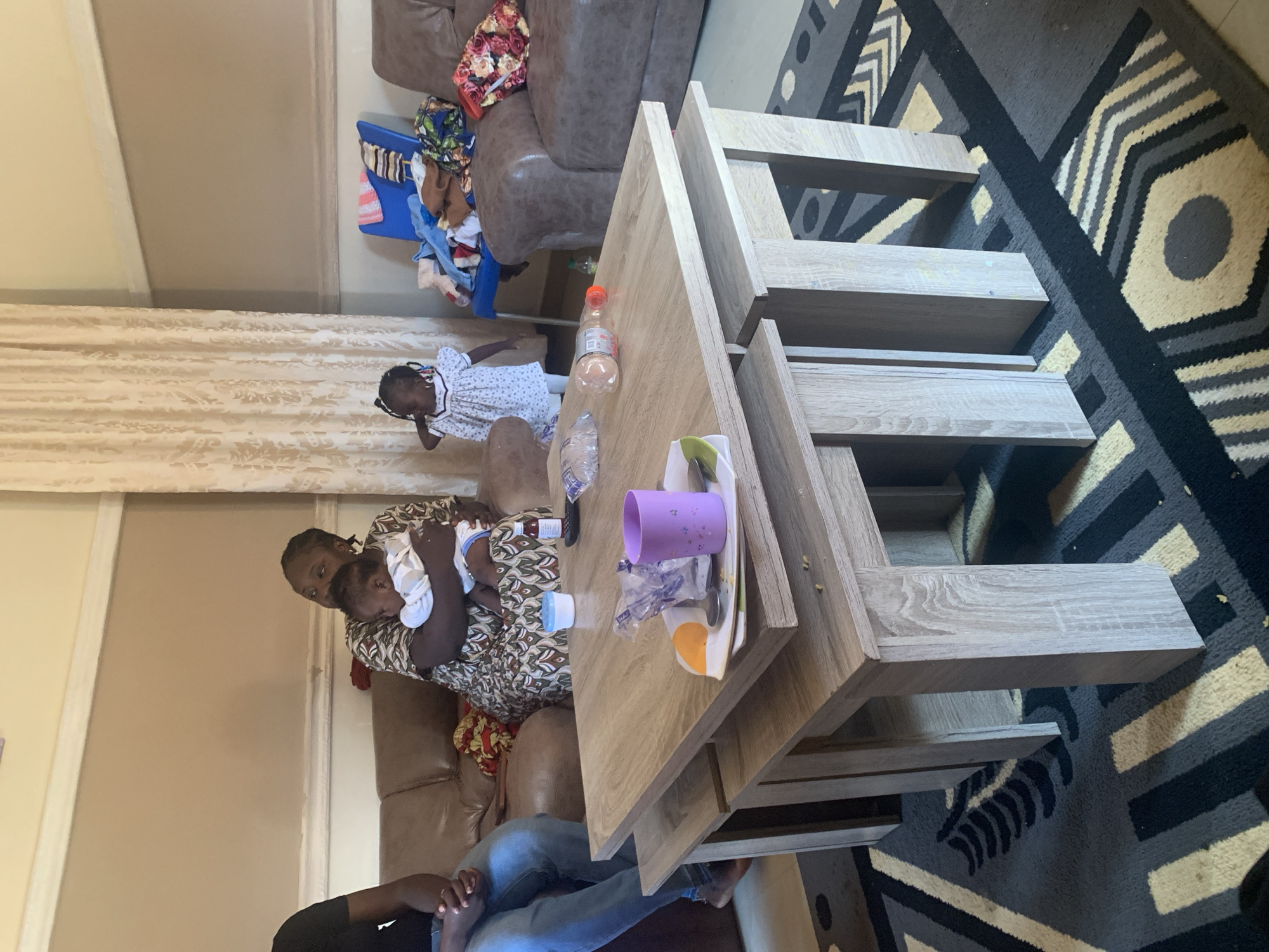 The image depicts an indoor scene, likely a living room, situated in Gura Top, Nigeria. In the foreground, a light-colored wooden nesting coffee table is prominent. On the table, there are several items including a purple cup, a plate with a spoon and some crumbs, a plastic bag with an orange lid, and a clear plastic bottle. To the left of the table, a person is seated, wearing blue jeans and a dark t-shirt, with their hands clasped.
Behind the table, a woman is seated on a brown leather sofa, holding a baby. The woman is wearing a patterned dress. Another young child, a girl in a white dress, stands near a cream-colored curtain. The background reveals more of the brown leather sofa, a blue plastic chair laden with clothes, and a patterned rug on the floor. The walls are a neutral beige color. The overall impression is of a domestic setting.