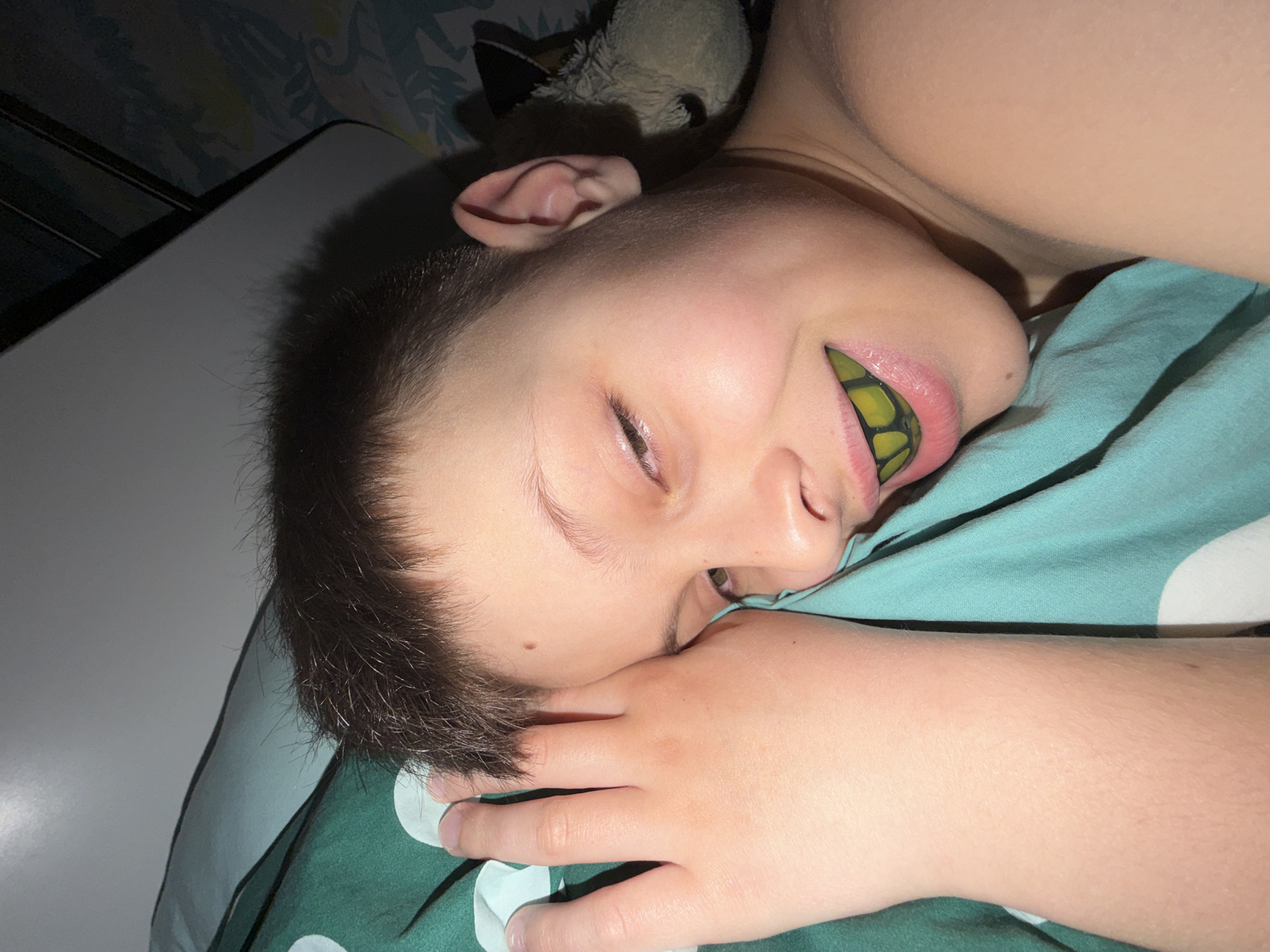 A child is lying on a bed. The bed has a green and white polka-dot patterned bedsheet. The child is resting their head on their hand, with a yellow and black patterned object in their mouth. A stuffed animal is visible near the child's head. A table is next to the bed. The location in the city of Votkinsk, Russia is not apparent.