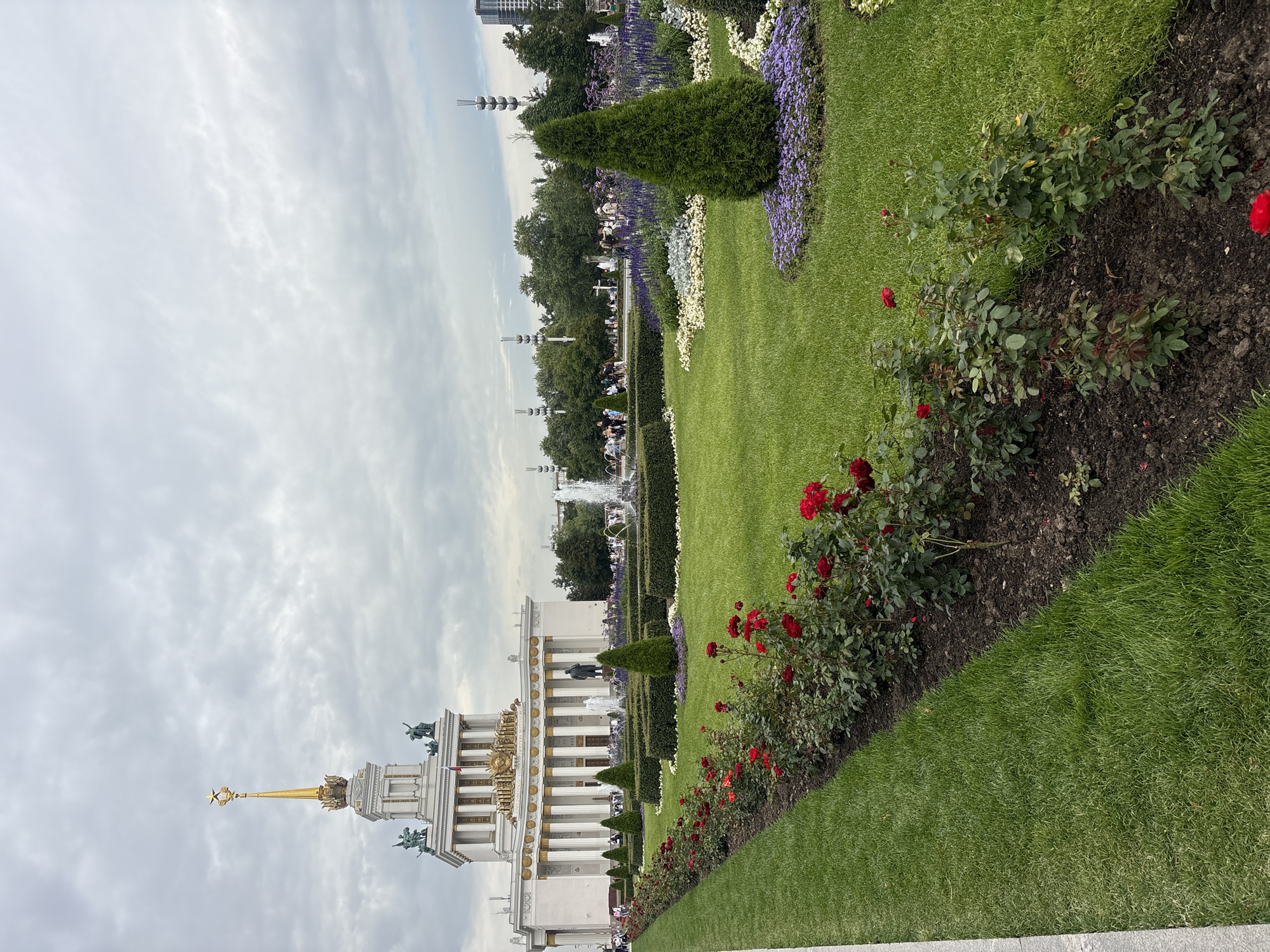 This is a medium shot of the All-Russian Exhibition Center (VDNKh) in Moscow, Russia, on a cloudy day. The main subject of the image is the iconic central pavilion, a white neoclassical building adorned with columns and sculptures, topped by a golden spire with a star. In front of the pavilion, a meticulously landscaped garden features a long row of red roses, manicured green hedges, and flower beds with purple and white flowers. Several conical evergreen trees are strategically placed within the garden. In the background, fountains are visible, along with numerous people strolling through the park. Trees and other structures, including a modern skyscraper, line the horizon. The overall scene depicts a popular public space with grand architecture and well-maintained greenery.