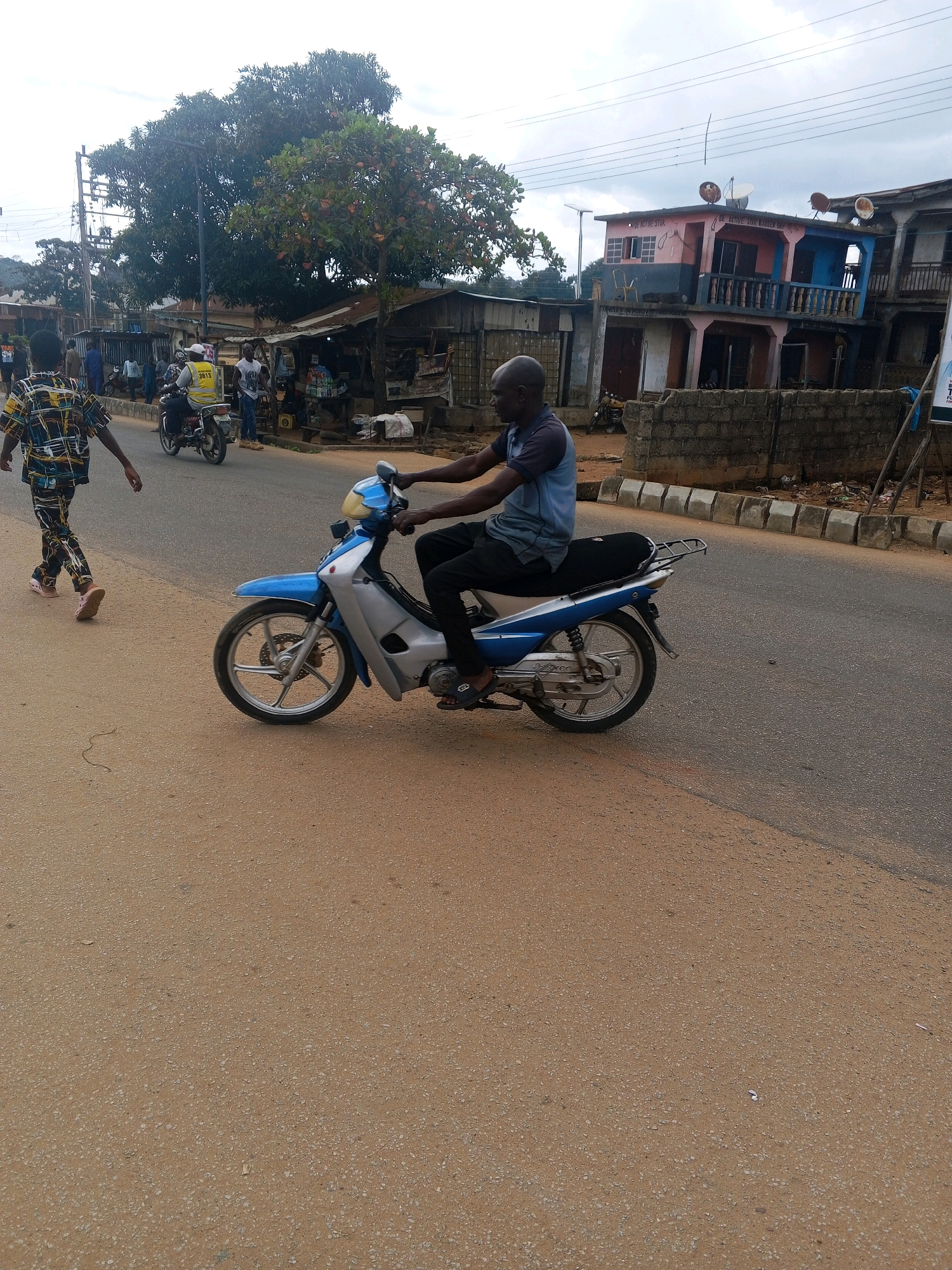A man is riding a blue and silver motorcycle on a paved road in Egbedi, Nigeria. He is wearing a blue t-shirt and black pants. The motorcycle is a 2016 model, and it has a silver rack on the back. The road is lined with buildings and trees. There are other people walking and riding motorcycles in the background. The sky is cloudy.