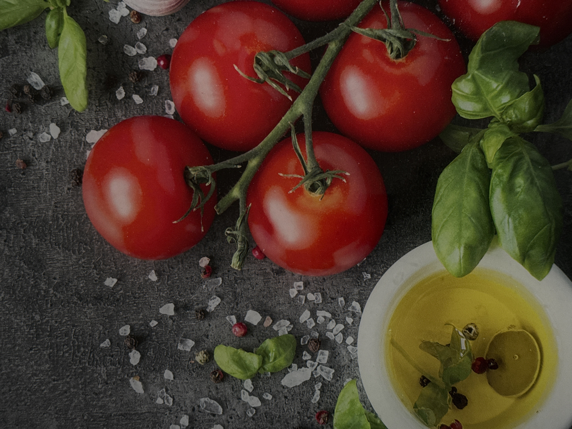 The image depicts a top-down view of fresh ingredients arranged on a dark, textured surface, likely a countertop or table. The primary subjects are several ripe, red tomatoes clustered on a vine. To the left, a small white bowl contains golden olive oil, with a few peppercorns and a sprig of basil floating within it. Several fresh basil leaves are scattered around the tomatoes and the bowl. Coarse salt crystals and whole black peppercorns are also visible on the dark surface. A hint of a garlic bulb is visible in the upper right corner. The lighting is dramatic, with highlights on the tomatoes and oil, creating a sense of depth and richness. There are no people or animals in the image, and no specific city or geographical location is identifiable.