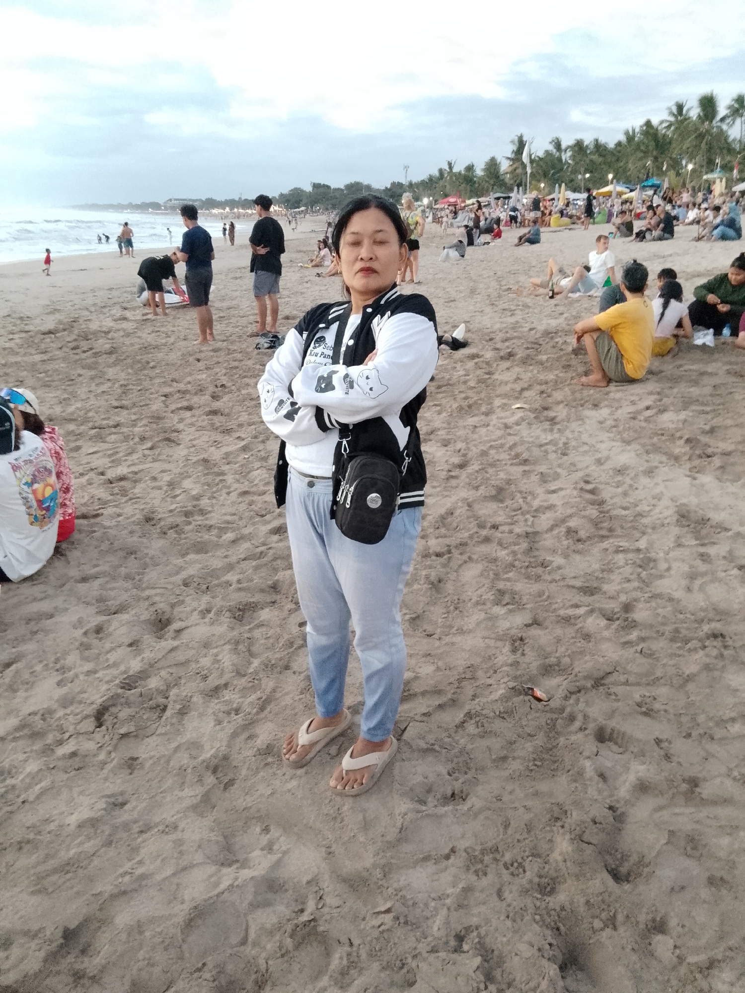 A medium shot captures a woman standing on a sandy beach in Kuta, Indonesia. She is positioned centrally in the frame, facing forward with her arms crossed and a neutral expression. She wears a white t-shirt, a black and white bomber jacket, light blue jeans, and beige flip-flops. A small black crossbody bag is slung over her right shoulder.
The background reveals a bustling beach scene with numerous people spread across the sand. To the left, the ocean meets the shore with gentle waves. Further back, a line of palm trees and umbrellas suggest a developed tourist area. The sky is overcast, indicating a cloudy day. Various individuals are engaged in activities like sitting, walking, and relaxing on the beach. The sand is a light brown color, showing footprints and some scattered debris.