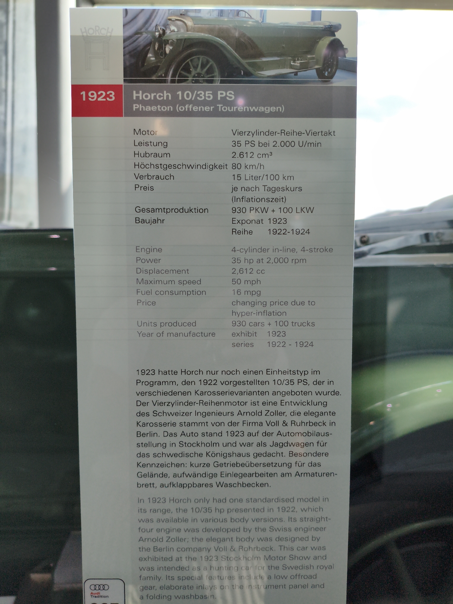 The image shows a display at the Audi Museum in Ingolstadt, Germany, featuring a 1923 Horch 10/35 PS Phaeton. The display includes a large photograph of the car and a descriptive plaque. The plaque is divided into sections, presenting technical specifications in both German and English, as well as a historical narrative about the car.
The German section lists details such as "Motor: Vierzyllinder-Reihe-Viertakt" (Engine: Four-cylinder in-line, four-stroke), "Leistung: 35 PS bei 2.000 U/min" (Power: 35 PS at 2,000 rpm), "Hubraum: 2.612 cm³" (Displacement: 2,612 cm³), and "Höchstgeschwindigkeit: 80 km/h" (Maximum speed: 80 km/h). Other German details include fuel consumption ("Verbrauch: 15 Liter/100 km"), price ("Preis: je nach Tageskurs (Inflationszeit)"), total production ("Gesamtproduktion: 930 PKW + 100 LKW"), and manufacturing year ("Baujahr: Exponat 1923, Reihe 1922-1924").
The English section mirrors these specifications, with "Engine: 4-cylinder in-line, 4-stroke", "Power: 35 hp at 2,000 rpm", "Displacement: 2,612 cc", and "Maximum speed: 50 mph". Fuel consumption is listed as "16 mpg" and price as "changing price due to hyper-inflation". "Units produced" are "930 cars + 100 trucks", and "Year of manufacture" is "exhibit 1923, series 1922 - 1924".
The descriptive text explains that in 1923, Horch had only one standardized model in its range, the 10/35 PS, which was available in various body versions. The car's four-cylinder engine was developed by Swiss engineer ArnoldZoller. The elegant body was designed by the Berlin company Voll & Ruhrbeck and was exhibited at the 1923 Stockholm Motor Show. It was intended as a hunting car for the Swedish royal family. Special features include a low off-road gear, elaborate inlays on the instrument panel, and a folding washbasin.
The photograph shows the front and side profile of a vintage green car, likely the Horch 10/35 PS Phaeton, positioned on a display platform. The environment appears to be an indoor museum setting, with controlled lighting and the presence of other displays or exhibits blurred in the background. The overall impression is one of historical preservation and public exhibition of automotive heritage.