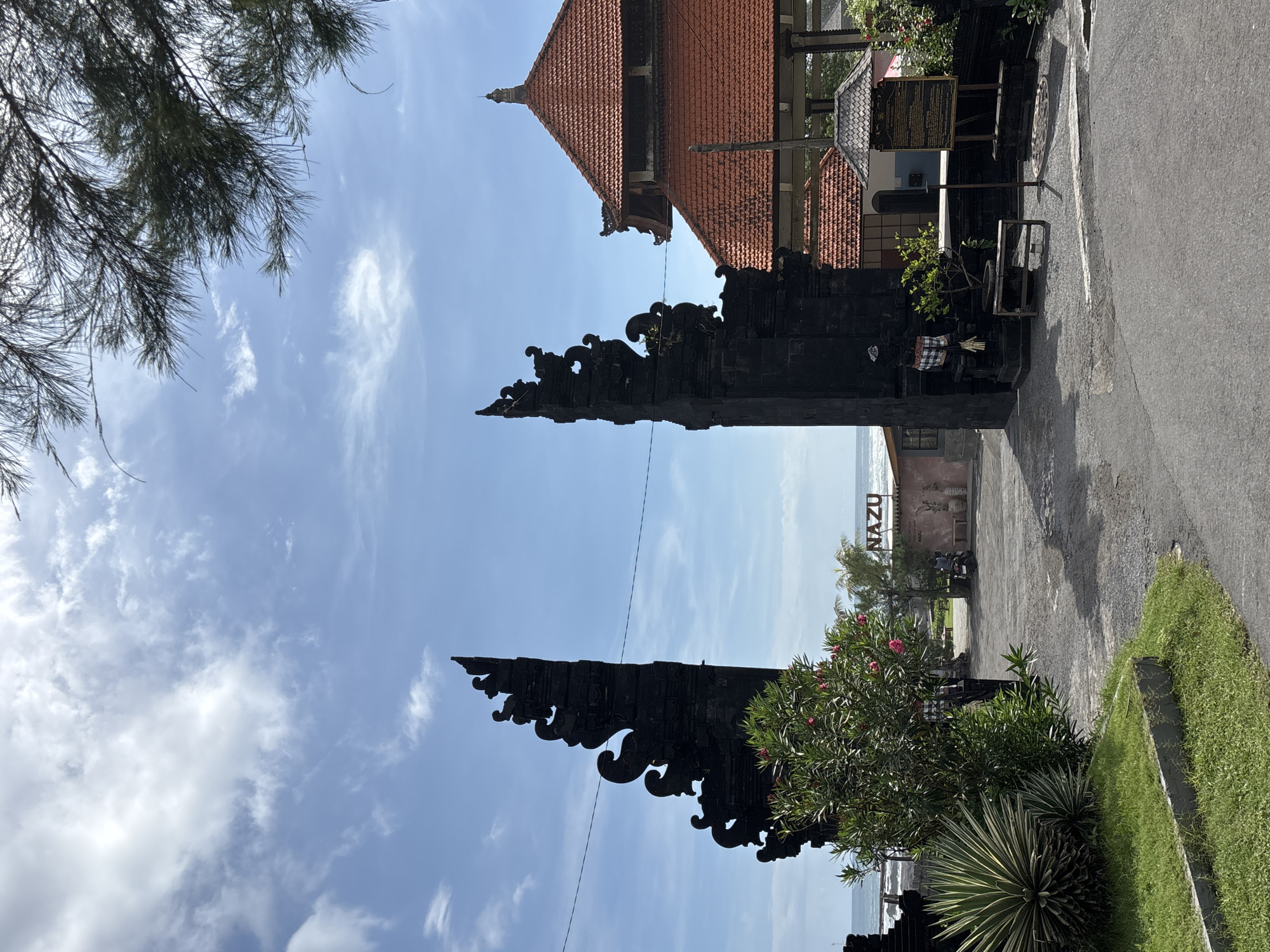 This is an exterior shot in Mengwi, Indonesia. A traditional Balinese stone gate with ornate carvings frames a view towards the coast. The gate is partially obscured by a tree with pink flowers on the right and some pine tree branches on the left. Beyond the gate, there is a concrete area with the word "NAZU" displayed in large letters. To the right of the concrete area is a paved road. The sky is blue with scattered clouds. In the background, part of a building with a red tile roof is visible.