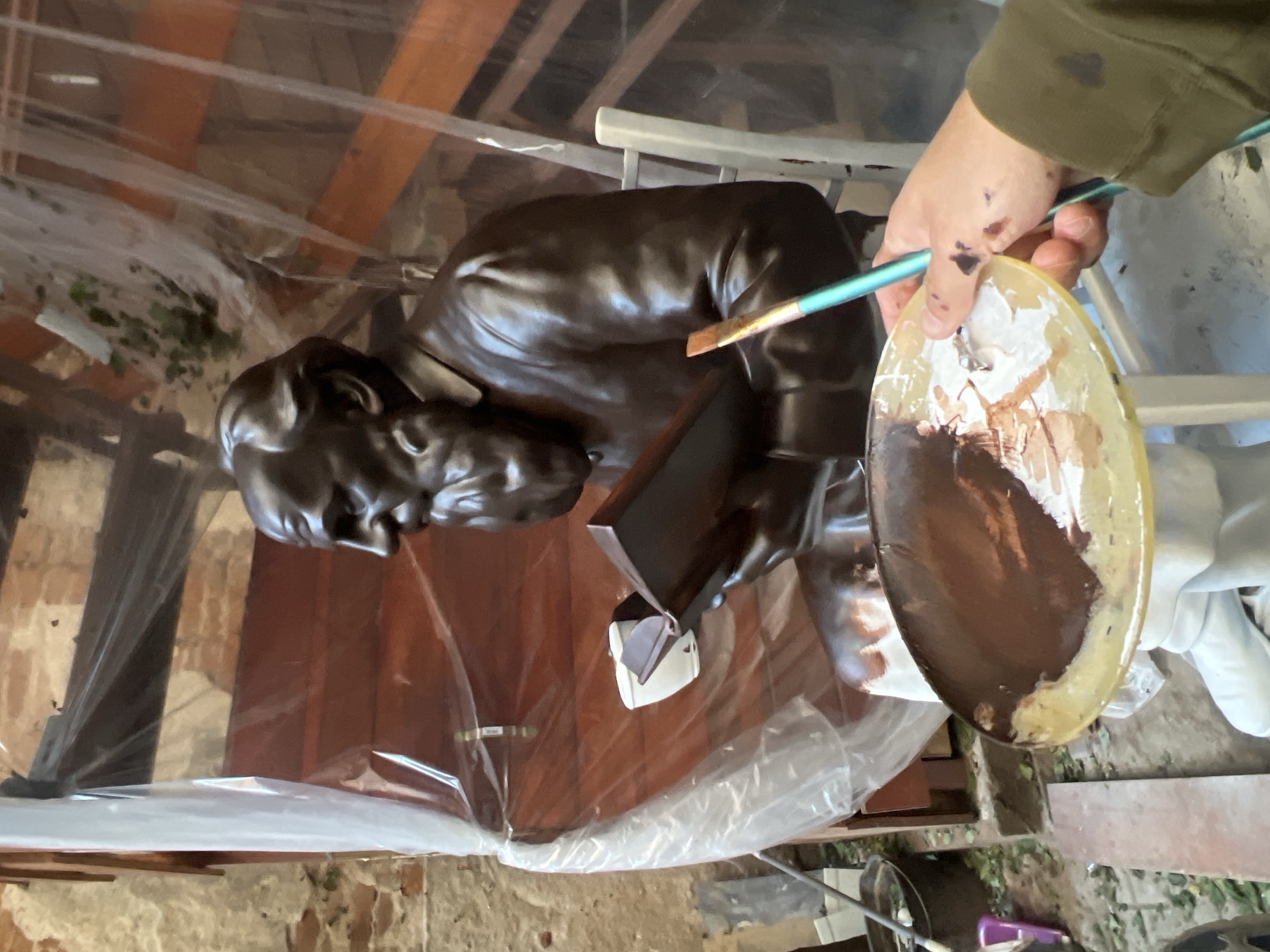 A person is painting a brown statue of an older man indoors. The statue appears to be placed in a space undergoing renovations or with exposed wooden beams in the ceiling. The person is holding a paint palette with brown and white paint and using a brush to apply paint to the statue. The statue is partially covered in plastic sheeting, and the scene appears to be set up for painting. The exact location within Plovdiv, Bulgaria is not identifiable from the image.