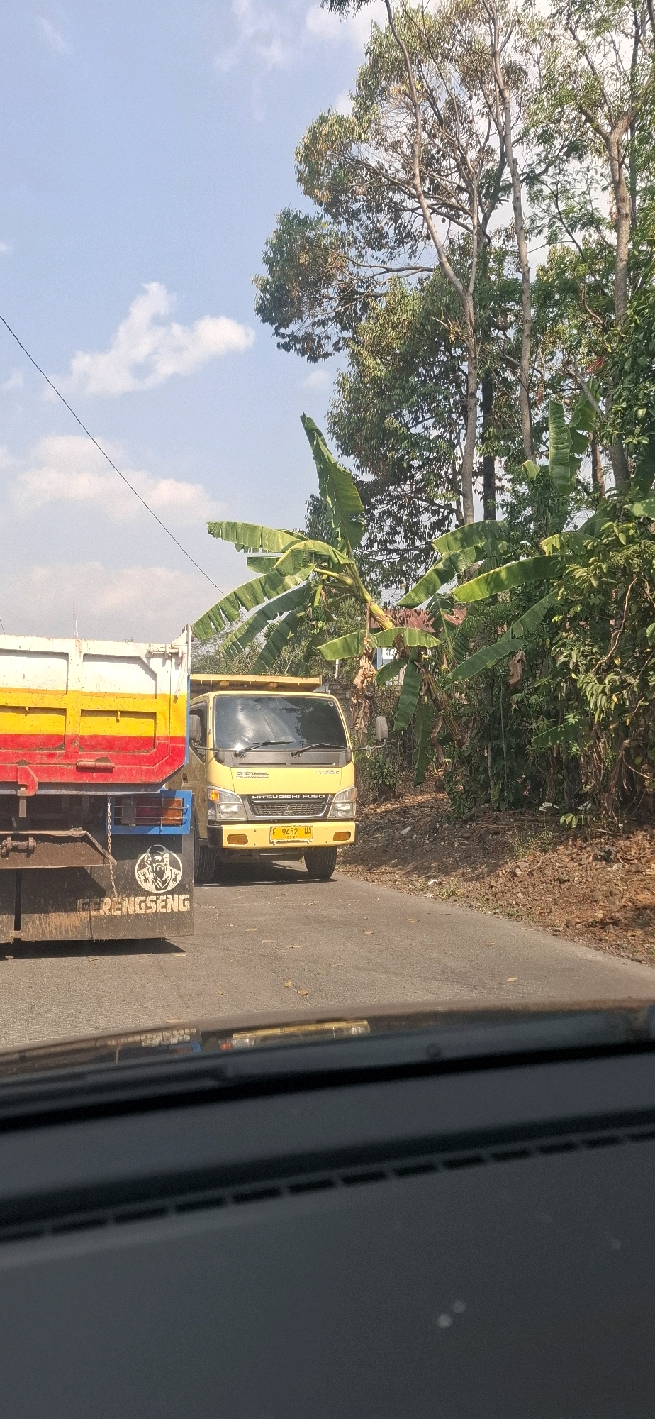 The image shows a road scene with two trucks and lush vegetation. In the foreground, the rear of a red, yellow, and white truck is visible, with the word "GERENGSENG" painted on its back. To the right, a yellow Mitsubishi Fuso truck is driving on the road. The truck has a license plate that reads "F 9452 WA". The surrounding area is filled with various trees and banana plants, suggesting a rural or semi-rural environment. The sky is blue with scattered clouds. The image appears to be taken from inside a vehicle, as evidenced by the dashboard reflection in the lower part of the frame. The location is specified as Sirnagalih, Indonesia.