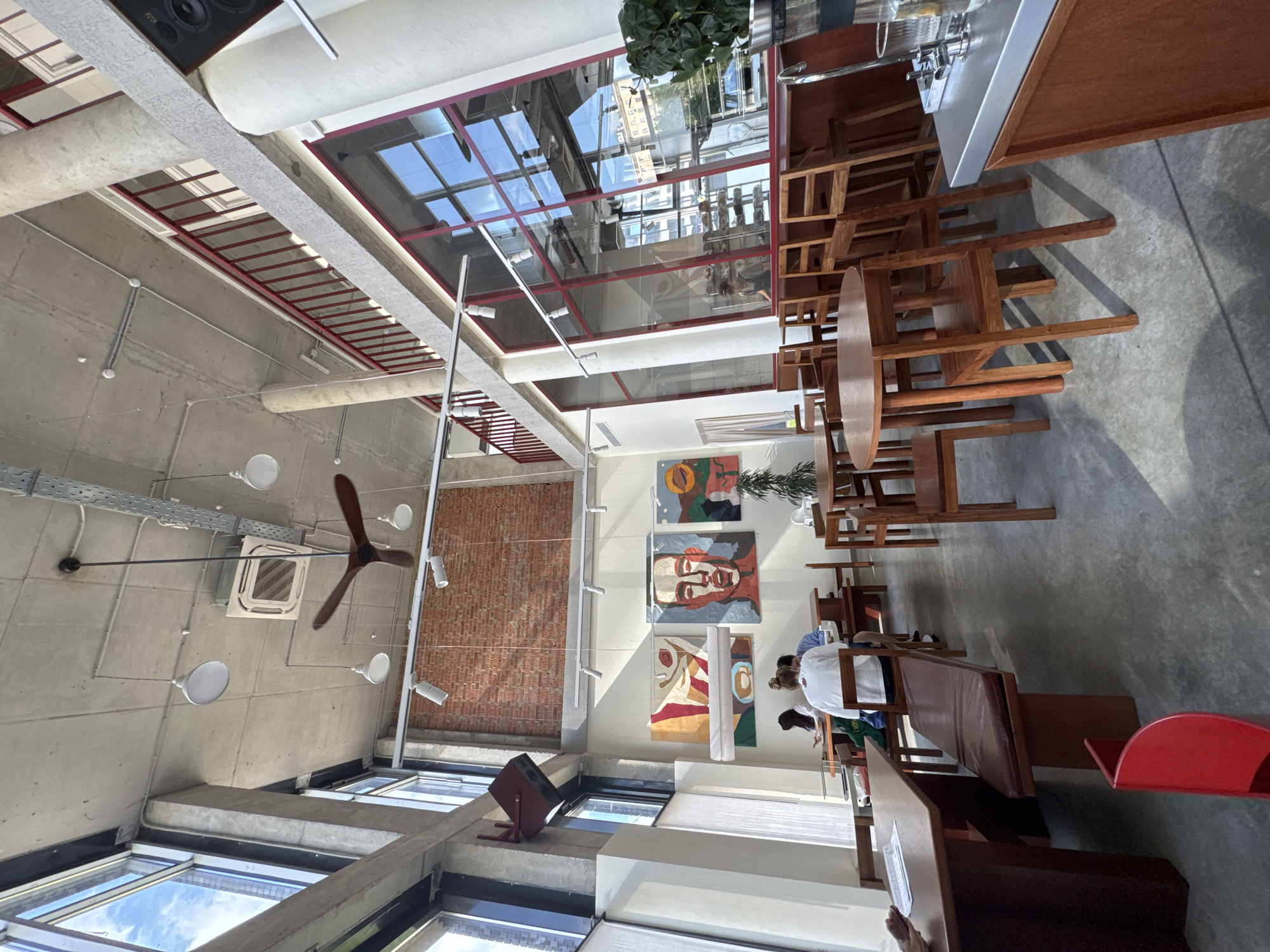 This is a medium shot, eye-level, interior photograph of a bright, open-concept cafe with a loft-like industrial design. The space features a polished concrete floor, exposed concrete ceilings, and large windows that let in ample natural light. Red accents are visible throughout the space, including a railing on an upper level, trim around the windows, and a modern red chair in the foreground.
The cafe is furnished with numerous wooden tables and chairs, including a long bench seat. Several people are seated at tables in the background, some of whom appear to be patrons and others possibly staff. Artwork, including abstract paintings and portraits, adorns the brick wall in the middle ground. Industrial elements such as exposed ductwork, lighting fixtures, and a large ceiling fan are prominent. A bar area with a stainless steel countertop and tap is visible on the right side of the frame, with a plant and glassware on display. The large windows on the left offer a view of the sky and city buildings, suggesting an urban setting. Based on the context of the image provided by the user, this cafe is located in Moscow, Russia.