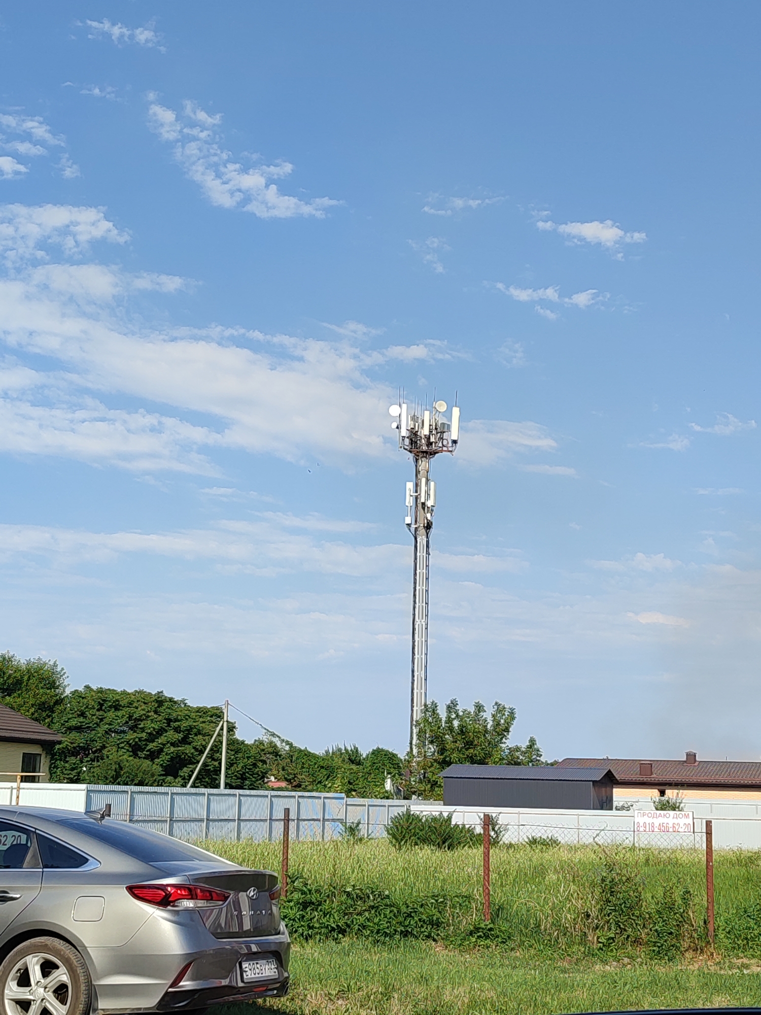 The image depicts a clear, blue sky with scattered white clouds, above a landscape that includes a tall telecommunications tower. The tower is a prominent structure, white in color, with multiple antenna arrays at its peak. Below the tower, a corrugated metal fence runs horizontally across the scene, separating a grassy area from a residential section. On the left, a gray sedan is parked, with its rear visible. To the right of the fence, a sign reads "ПРОДАЮ ДОМ" (FOR SALE HOUSE) with a phone number. In the background, houses with brown and yellow roofs and trees are visible. The scene is set in the city of Abinsk, Russia.