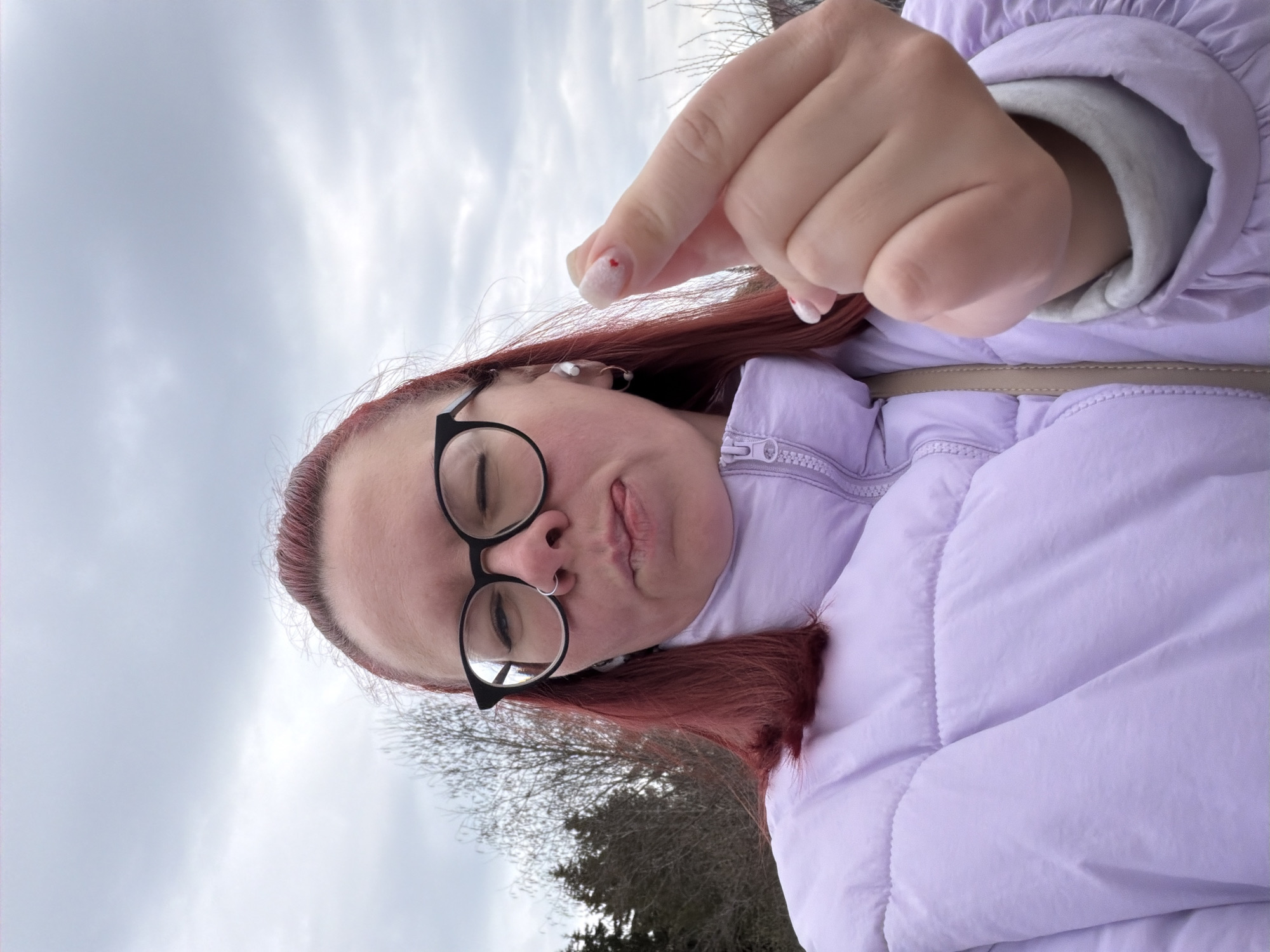 A person with reddish-brown hair and black-rimmed glasses is shown in a close-up, slightly tilted shot. They are wearing a light purple puffer jacket over a grey collared shirt. Their right hand is raised, with the index finger extended towards the camera, showing a white nail with a red heart design. The person appears to be wearing a nose ring. The background shows bare trees and a cloudy sky. The location is not explicitly evident from the image to confirm it's Chaikovsky, Russia.