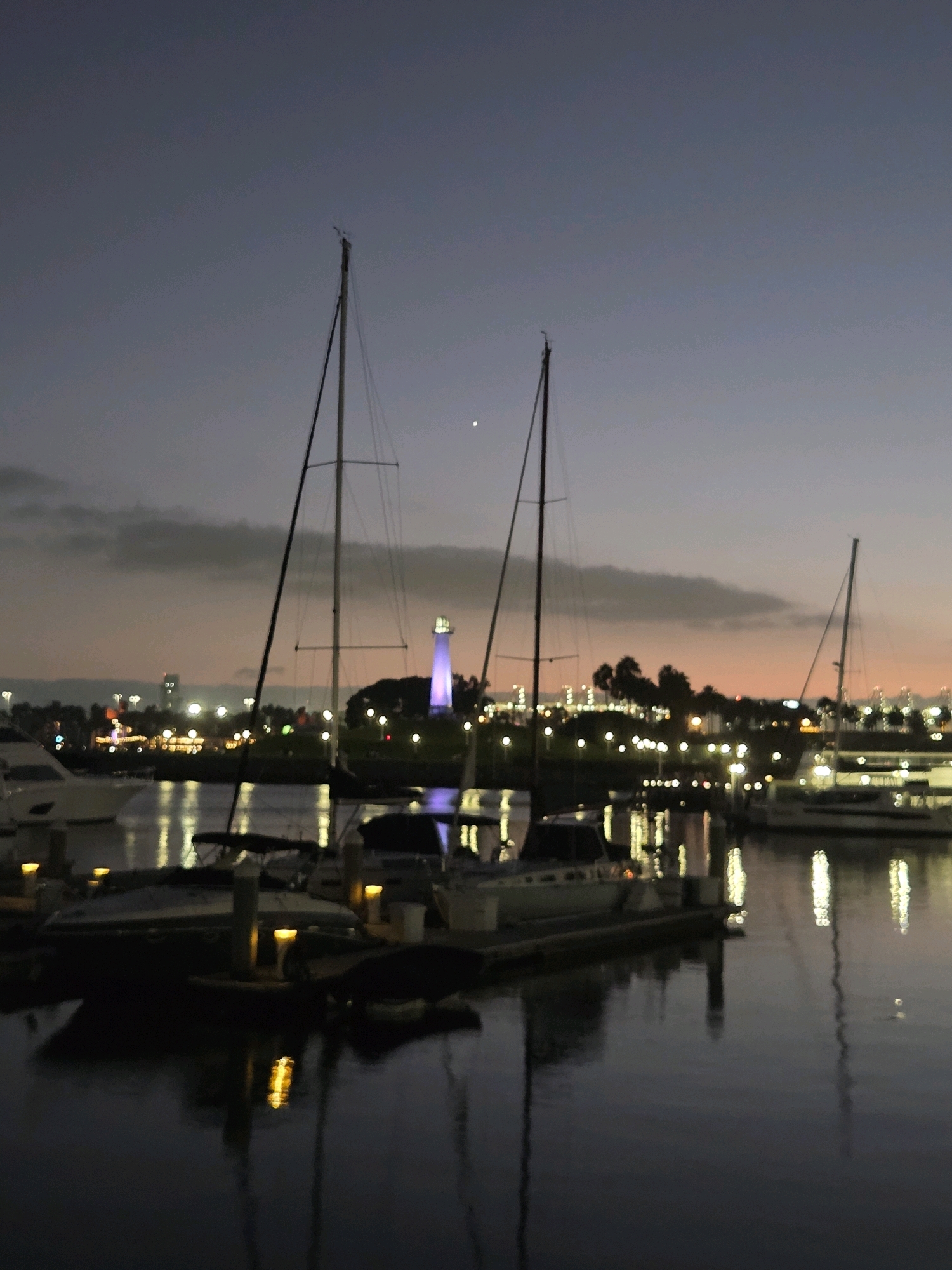 The image depicts a waterfront scene in Long Beach, California, at dusk. Several sailboats are docked in a marina, their masts silhouetted against a twilight sky. In the background, a purple lighthouse stands prominently, surrounded by city lights and palm trees. The water reflects the lights from the shore and the boats, creating a shimmering effect. The overall atmosphere is calm and serene, with the last vestiges of daylight fading into the night.
