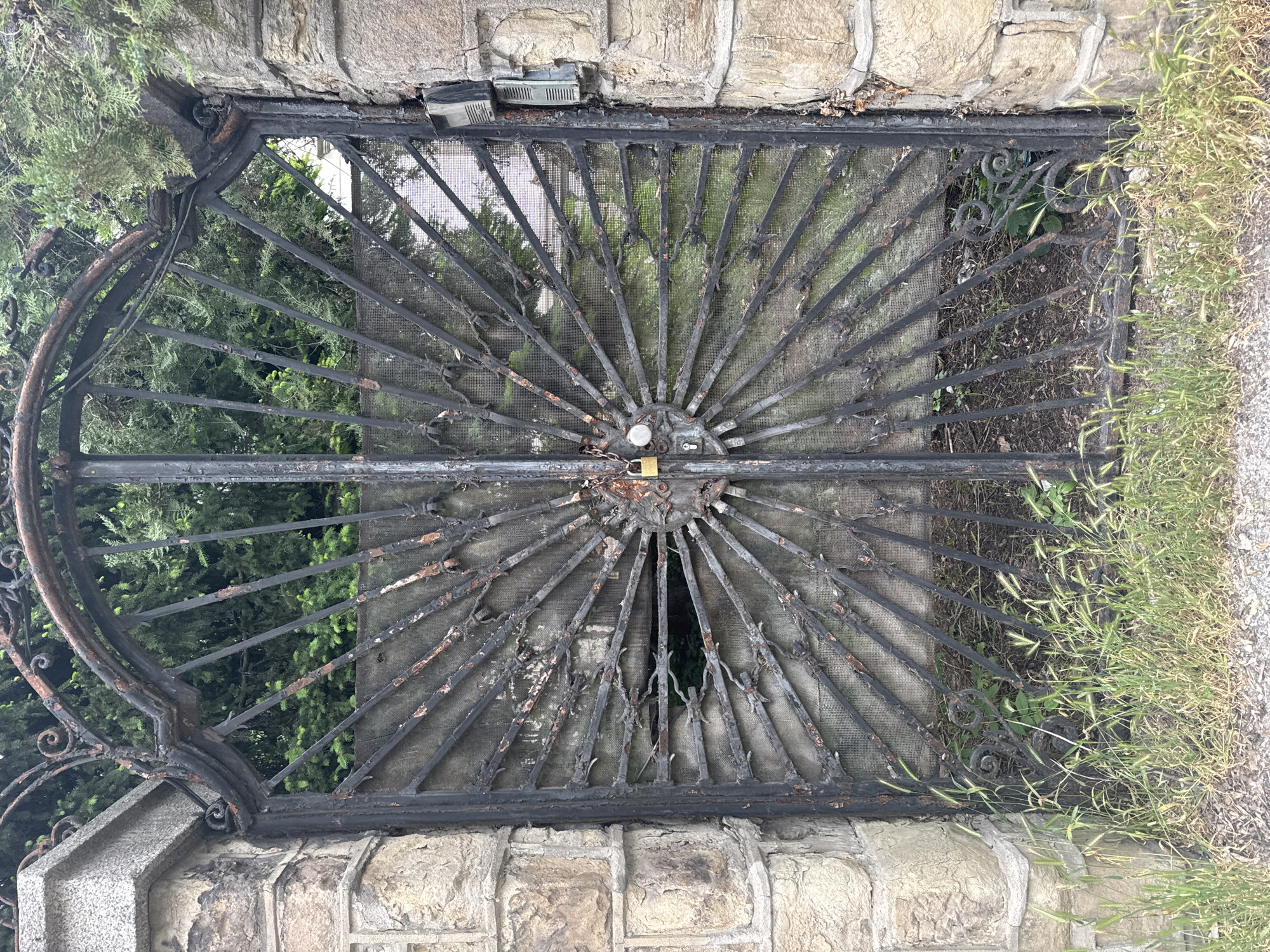 An eye-level, full shot depicts a weathered, arched, black metal gate set into a stone wall. The gate is divided into two halves that meet in the center, secured with a golden padlock and a chain. The gate's design features a sunburst pattern with radiating metal bars and decorative metal scrolls at the outer edges. The metal is peeling and shows signs of rust. Behind the gate, vegetation is visible. The stone wall is composed of tan-colored stones. The scene is overgrown with weeds and grass along the edges. Given the architectural style of the stone and the presence of an arched iron gate, it is likely that this image was taken in Sofia, Bulgaria.