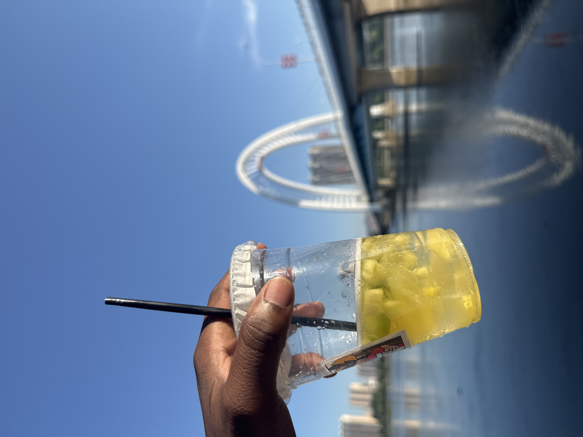 A close-up, eye-level shot shows a hand holding a clear plastic cup filled with a yellow, translucent beverage containing ice and fruit pieces. A black straw extends from the cup, and a white paper lid is partially visible. The cup has a colorful sticker on its side. In the background, out of focus, is a large, modern bridge with an arched structure spanning across a body of water. The sky is clear and blue. Buildings can be seen in the distance on the left side of the image. This appears to be a scene in the city of Linghe, China, given the distinctive architecture of the bridge.