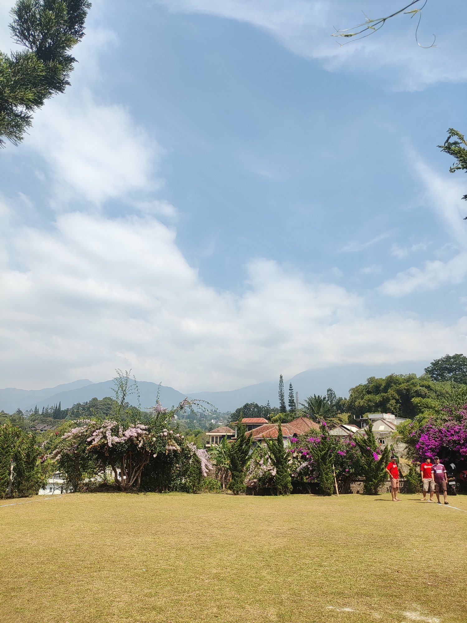 The image depicts a spacious, manicured lawn in the foreground, with a bright blue sky dotted with scattered white clouds overhead. In the midground, a line of lush green trees and vibrant purple flowers form a dense border, separating the lawn from a collection of residential buildings and a distant, hazy mountain range. Three men are visible standing on the lawn, two of whom are facing the camera and appear to be casually conversing. The overall setting suggests a pleasant, outdoor environment, likely a park or a residential complex with ample green space. The location is identified as Cisarua, Indonesia.