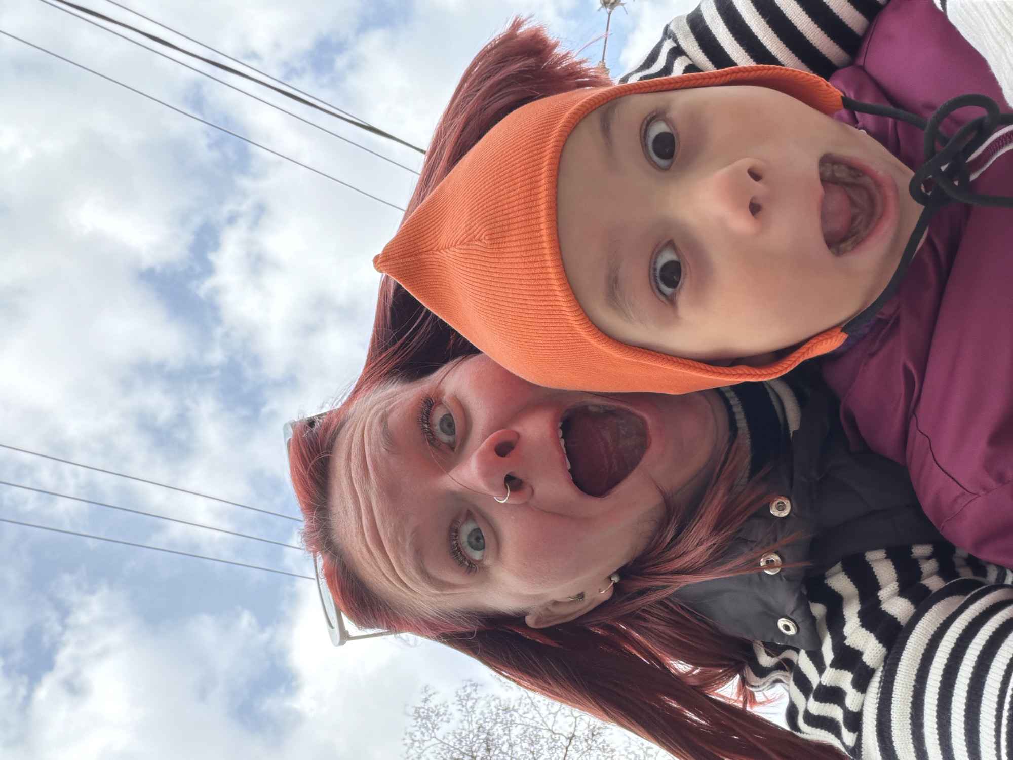 The image captures a playful, low-angle selfie featuring an adult and a young child looking upwards into the camera against a cloudy sky. The photo is rotated, giving a unique perspective.
In the foreground, an adult with reddish-brown hair, light-colored eyes, and a silver septum piercing, stares directly at the camera with wide, exaggerated eyes and an open mouth, as if screaming or expressing surprise. She wears a black and white striped top layered under a black vest.
Positioned above and slightly to the right of the adult is a child, mirroring the adult's expression with wide eyes and an open mouth. The child is wearing an orange pointed beanie and a purple padded jacket, also over a black and white striped garment.
The background consists of a bright, overcast sky with several thin utility wires stretching across the upper portion of the frame. Faint, bare tree branches are visible towards the bottom right, suggesting an outdoor setting during a cooler season. The overall scene conveys a moment of fun and exaggerated playfulness between the two individuals. There is no visible text. The location context is Chaikovsky, Russia.
