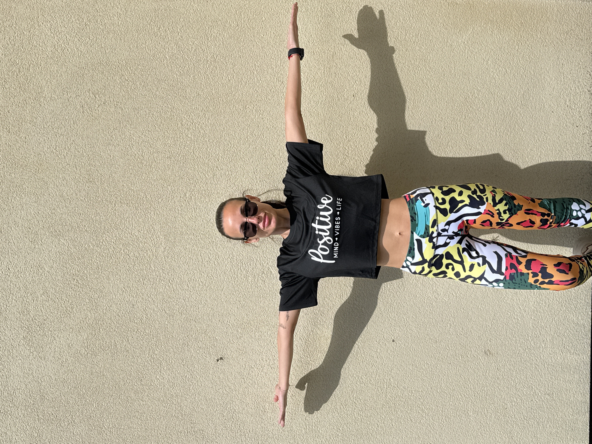 A person is standing facing forward, rotated ninety degrees counterclockwise, against a light-colored stucco wall. The person wears sunglasses and a black crop top with the word "Positive" printed on it, along with the phrase "MIND + VIBES + LIFE". She also wears patterned leggings. Her arms are outstretched perpendicular to her body. The background is a plain stucco wall. It is day. The location is the city of Pleven, Bulgaria.
