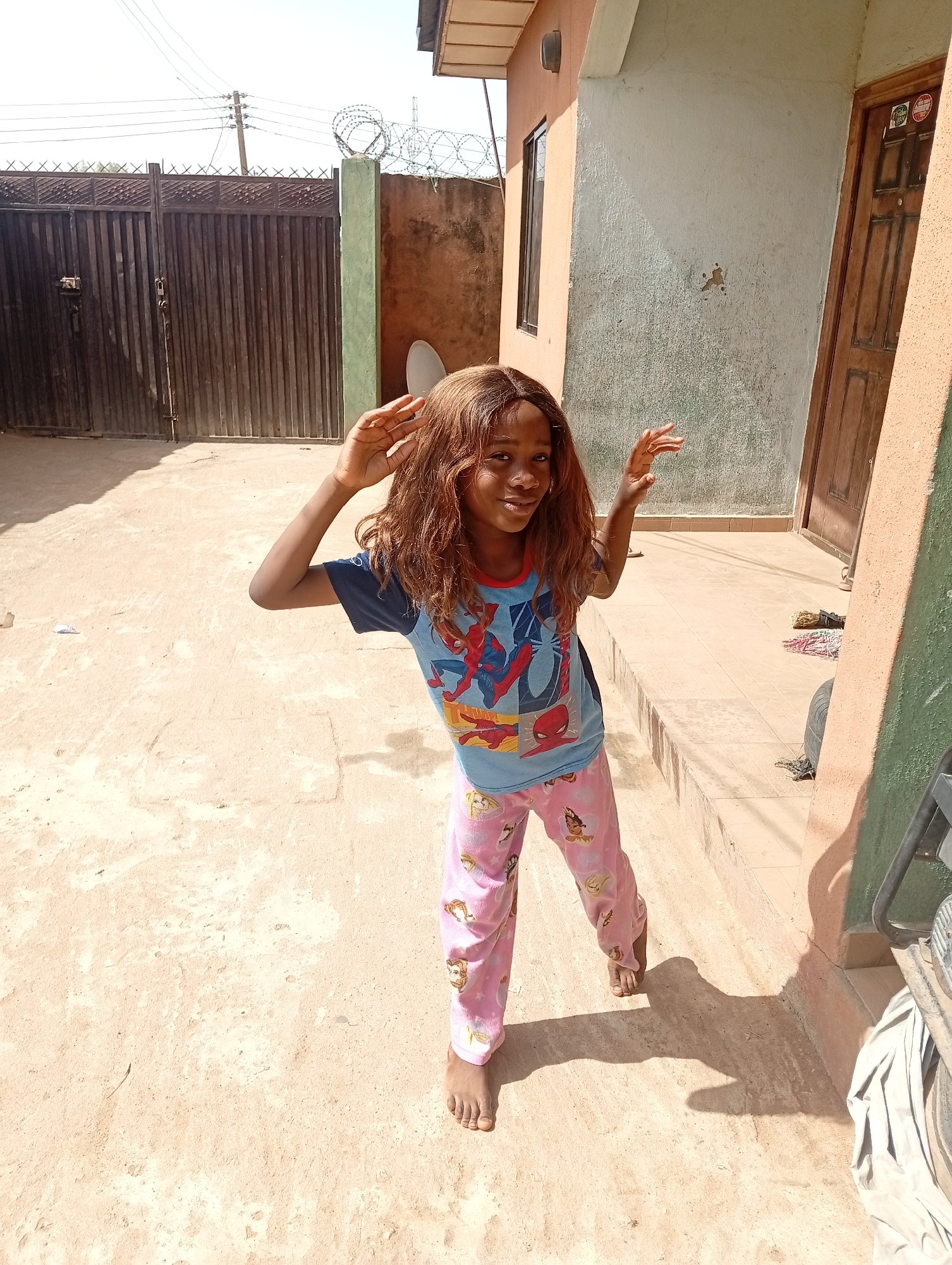 A young, barefoot, Black girl with long brown hair, wearing a blue Spiderman T-shirt and pink patterned pajama pants, stands in a sandy courtyard. She is smiling and has her hands raised near her head. Behind her is a dark metal gate, a light-colored building with a brown door, and a concrete courtyard. The location appears to be Sabon Tasha, Nigeria, given the typical building styles and overall environment. There is barbed wire atop the walls surrounding the property.