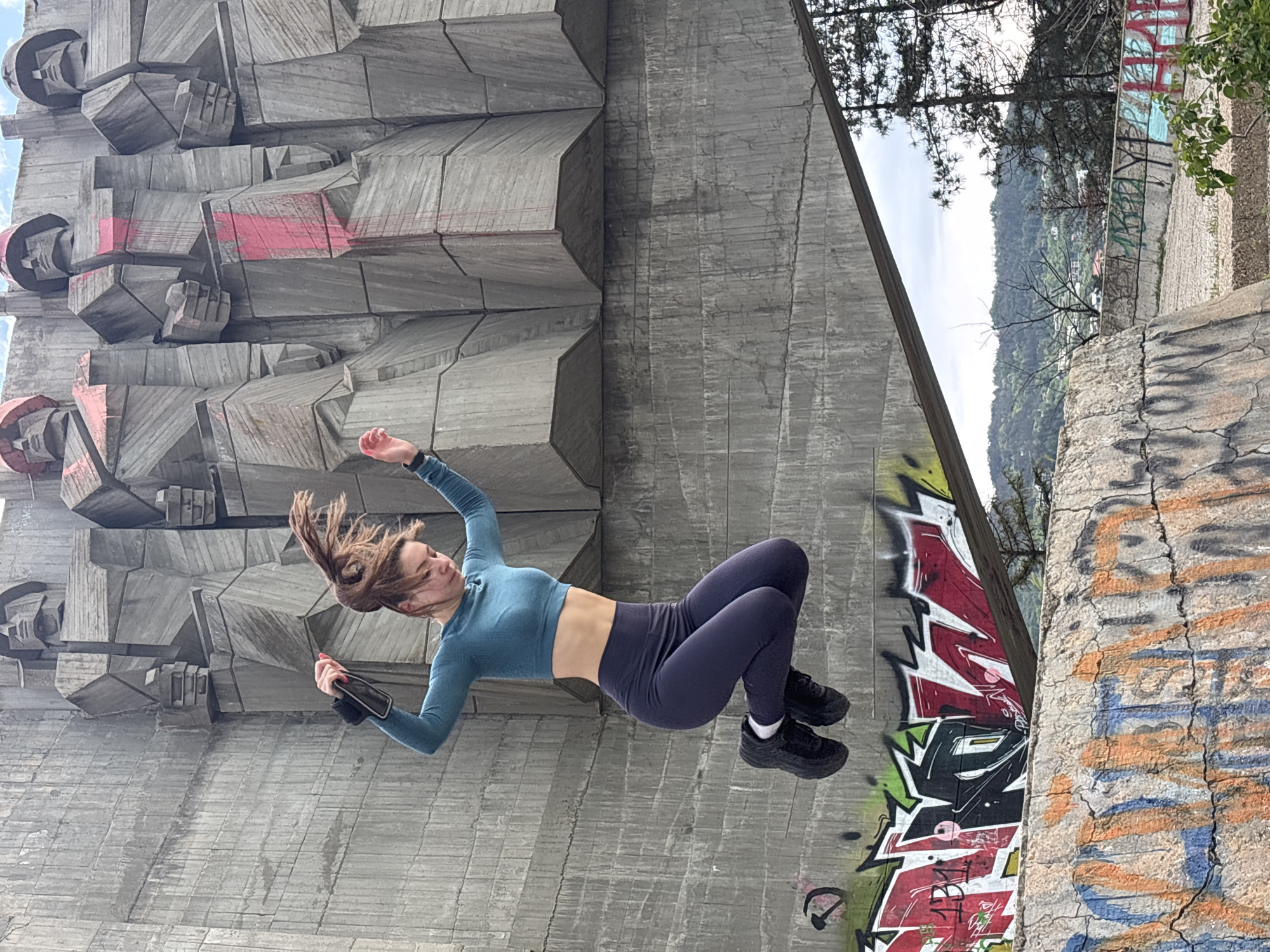 A person in athletic clothing is captured mid-air in what appears to be a dynamic action, perhaps a jump or flip. She's wearing a long-sleeved teal crop top and dark-colored athletic leggings, with black athletic shoes. Her hair is in motion, suggesting swift movement. In her left hand, she is holding a black device.
The background consists of two distinct areas. To the left and partially behind the person is a large, concrete structure composed of geometric shapes and Soviet-era sculptures. This is the Monument of the Bulgarian-Soviet Friendship, located in the city of Varna, Bulgaria. The structure has traces of red paint.
To the right, there's a concrete wall covered in graffiti, separating it from foliage and a view of the terrain beyond. The graffiti appears in shades of red, black, white, green, and grey. The sky is overcast.
