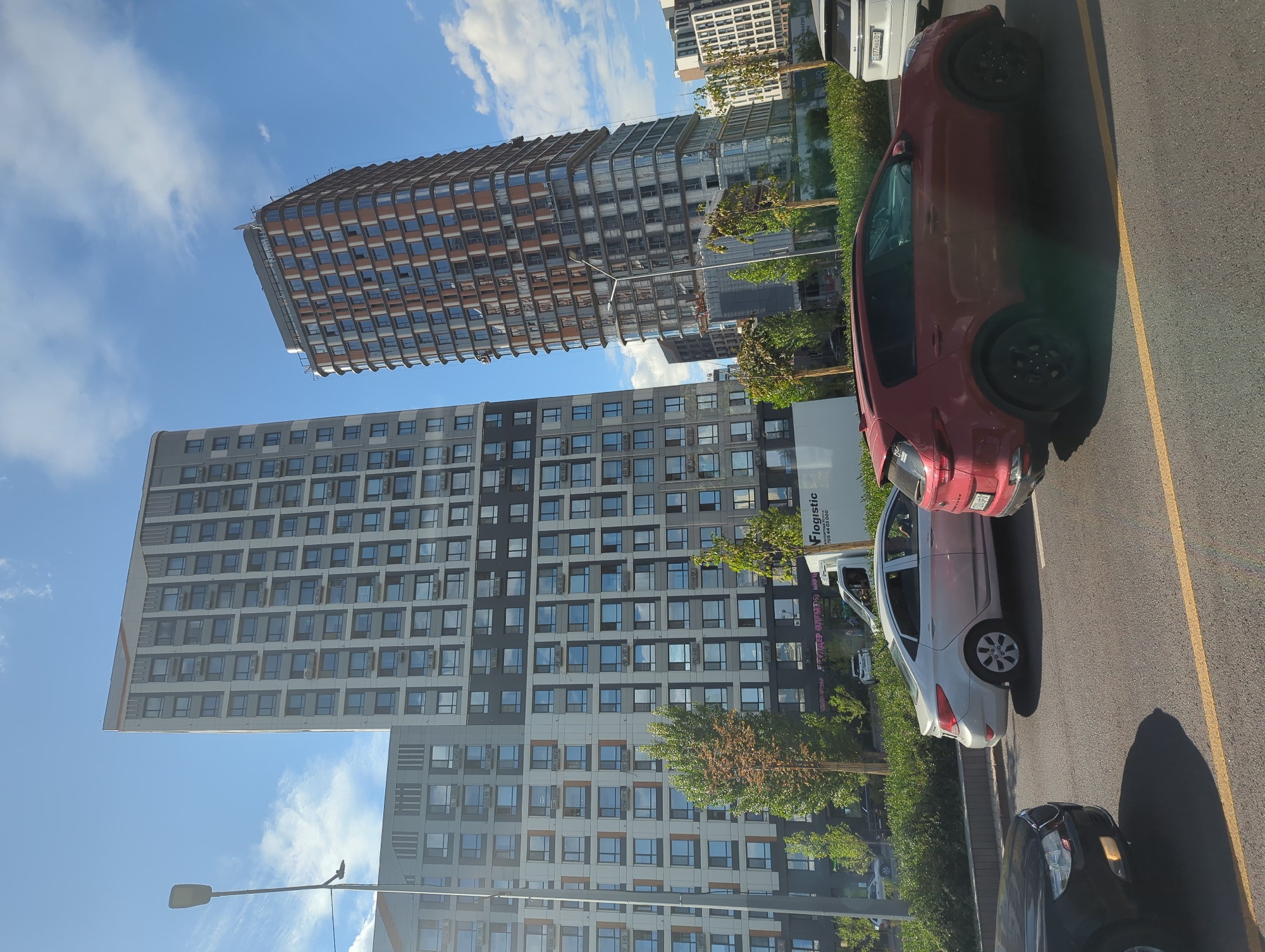 The image captures a street view in Astana, Kazakhstan, on a sunny day with scattered clouds. Two tall, modern buildings dominate the background, one appearing to be under construction with visible scaffolding and cranes. These high-rise structures feature a grid-like pattern of windows. In the foreground, several cars are parked or driving on a paved road. A red SUV and a silver sedan are most prominent, with a black car partially visible on the left. Green trees and bushes line the sidewalk area between the road and the buildings. A white delivery truck with "Flogistic" branding is parked near the buildings. The overall scene depicts an urban environment in Astana with ongoing development.