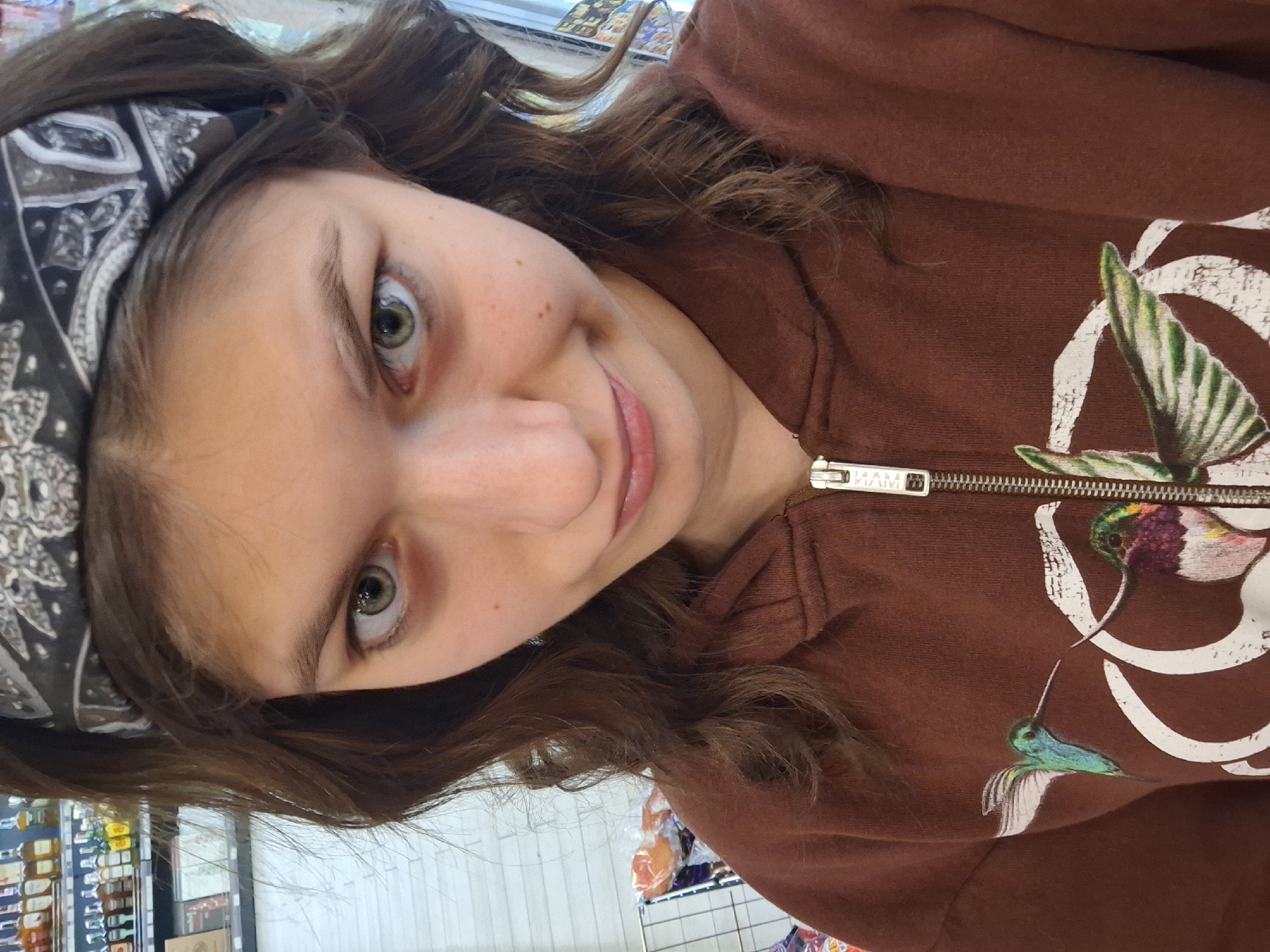 A young, fair-skinned person with brown hair and green eyes wearing a gray and black patterned headband and a brown zippered hoodie with hummingbird designs is pictured in a selfie. The person is smiling slightly. In the background, shelves stocked with various products are visible, suggesting the location is likely a store. Without further information or context, it's not possible to confirm if this photo was taken in Panagyurishte, Bulgaria.