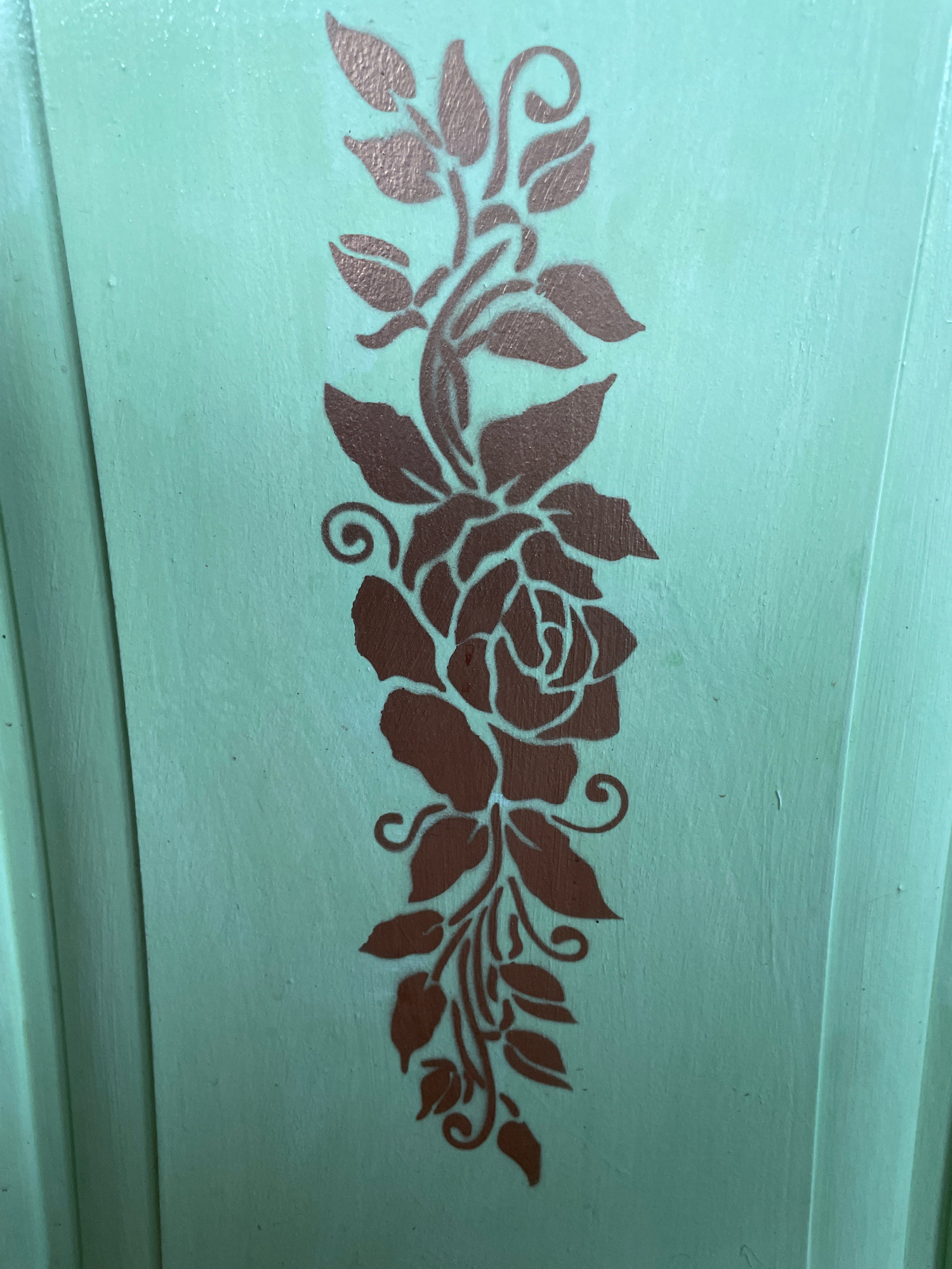 The image shows a close-up view of a decorative stencil of a rose vine against a mint green background. The stencil is a metallic copper color and features a central rose with detailed petals, surrounded by leaves and swirling tendrils. The background appears to be a painted surface, possibly wood or furniture, with a slightly textured finish. Vertical lines are visible on the right side of the image, suggesting the edge of a panel or a fluted design element. The lighting is even, highlighting the detail of the stencil. There are no people or any indication of a specific location within a city.