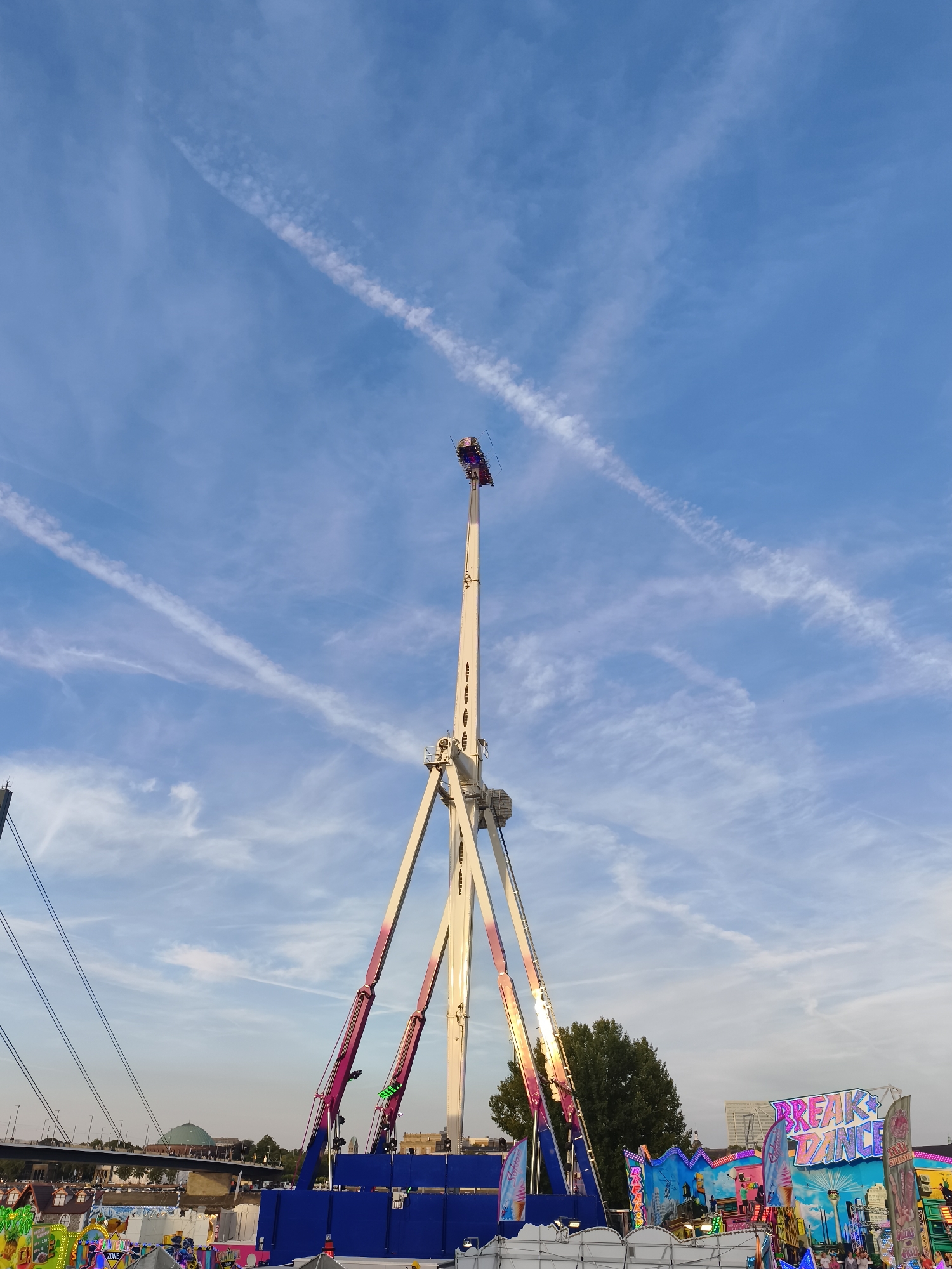 An amusement park ride, possibly a pendulum ride, dominates the center of the frame against a bright blue sky with wispy clouds. The ride's tall, cream-colored tower is supported by three angled legs, one of which is a bright pink and purple. A gondola, with people visible within it, is suspended at the very top of the tower. In the background, a bridge with traffic spans a body of water. To the right, colorful amusement park stalls, one with a sign reading "BREAK DANCE," are visible, with a few people milling about. Trees and buildings, including a dome-shaped structure, are discernible behind the stalls. The scene appears to be in Dusseldorf, Germany, possibly at the Rheinkirmes fair.