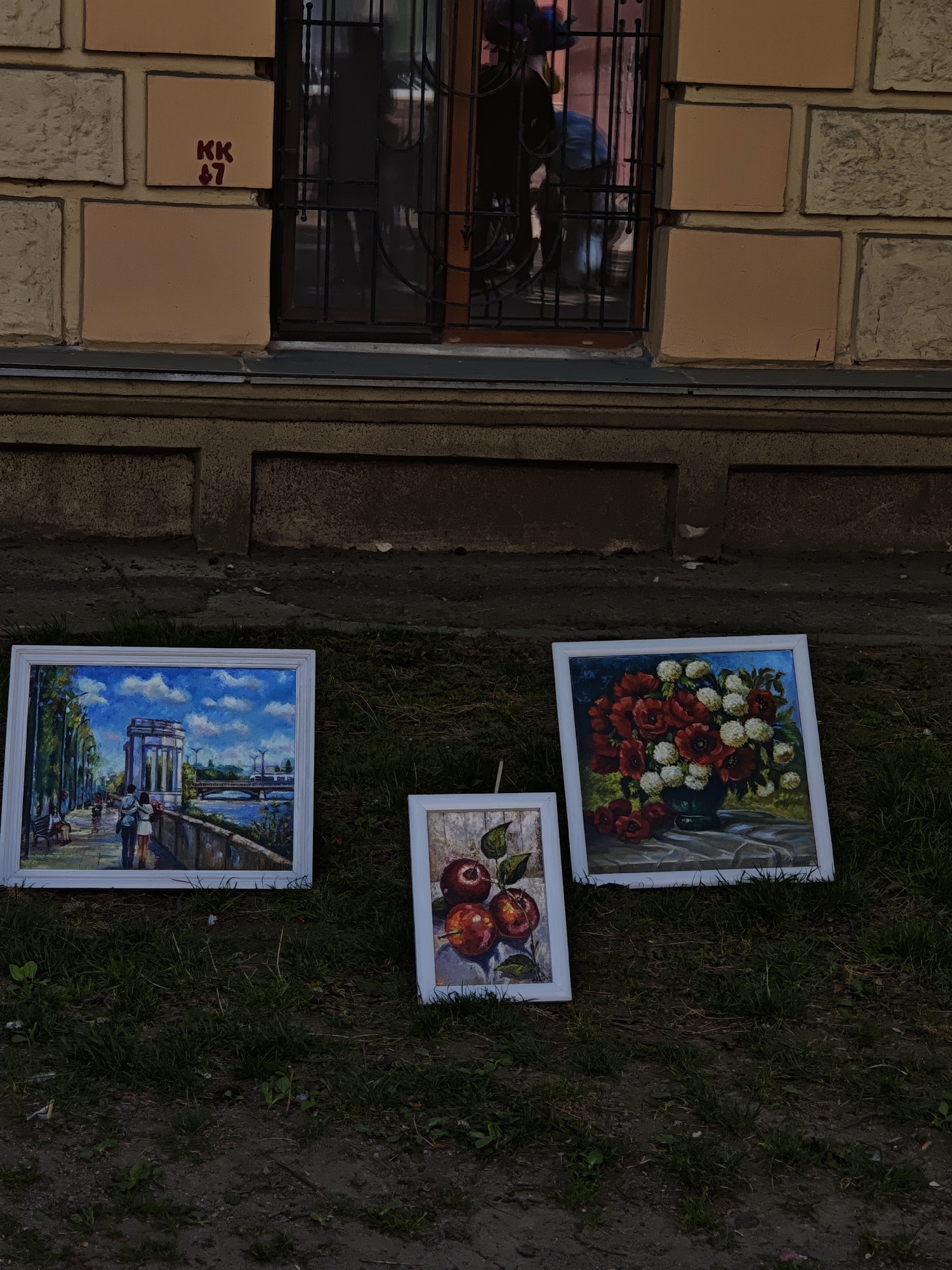 The image shows three framed paintings placed on a patch of grass in front of a building. The building's lower facade is composed of stacked blocks. Above, there's a window with a dark metal grate and a brick wall with “KK 67” written in red.
The left painting depicts a cityscape scene with a couple walking alongside a body of water, likely the Southern Bug river. The center painting portrays a still life of apples. The right painting features a vase filled with red and white flowers.
The scene is located in the city of Vinnytsia, Ukraine, indicated by "KK 67", which refers to the location of "Kotsiubynskoho Street, 67" in the city. No people are visible in the environment except in the paintings. The grass is green.