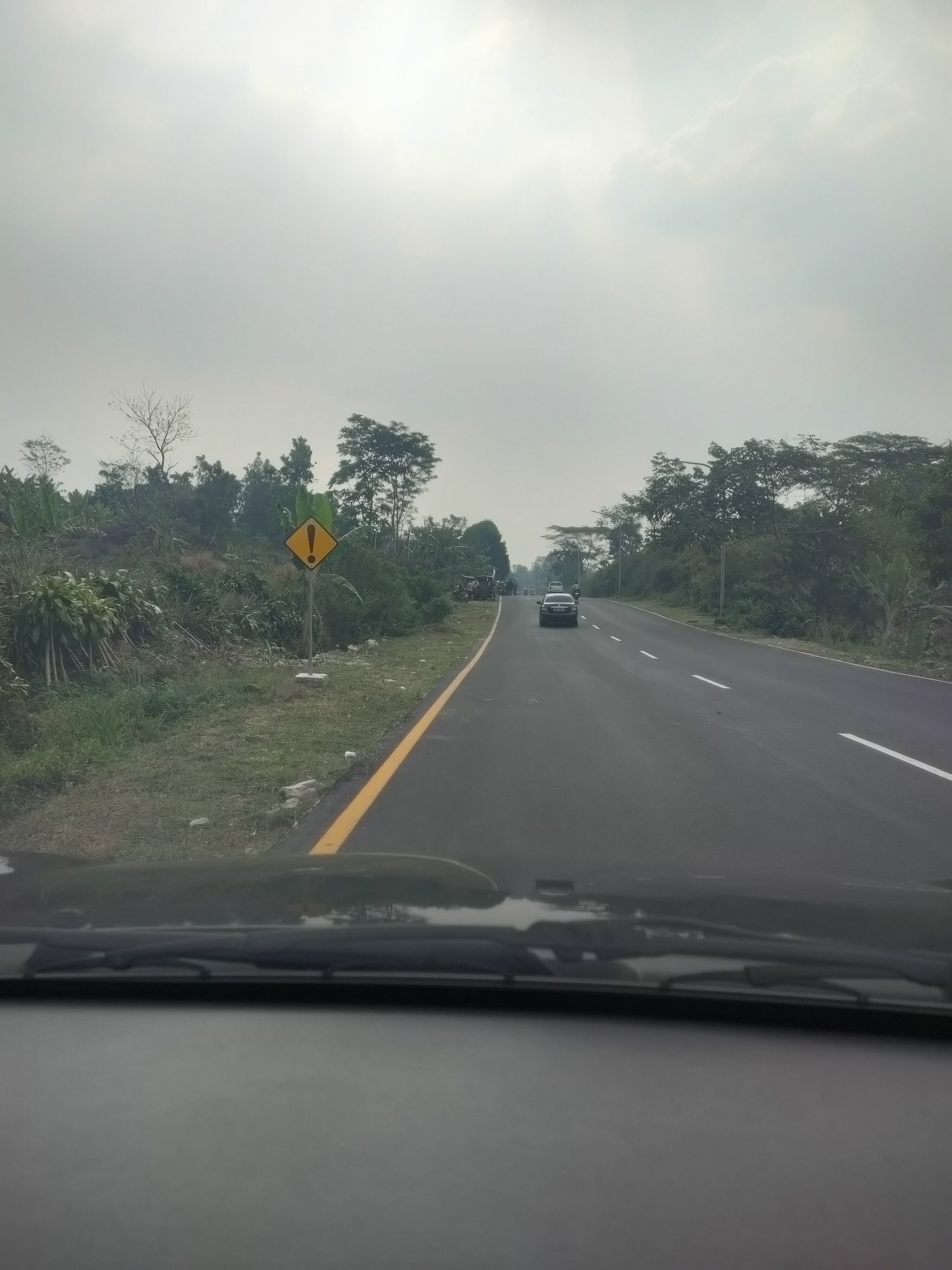 The image shows a two-lane asphalt road with a yellow line marking the edge on the right. The road is flanked by lush green vegetation and trees on both sides. A yellow diamond-shaped warning sign with a black exclamation mark is visible on the left side of the road. There are at least two cars on the road, one black sedan driving in the same direction as the camera and another white car further ahead. The sky is overcast and appears to be a pale gray. The perspective is from inside a vehicle, looking through the windshield, which is partially visible at the bottom of the frame. The scene depicts a rural or semi-rural road, possibly in the city of Subang, Indonesia, given the context provided.