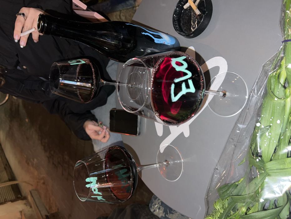 Three wine glasses sit atop a gray table, each filled with red wine and displaying names written in turquoise: "Mark," "Eva," and an obscured third. A dark wine bottle with a light blue design and an ashtray with cigarette butts are also visible. A hand holding a cigarette and a mobile phone are on the side, while a bouquet of flowers, wrapped in plastic, is placed near the edge of the table.