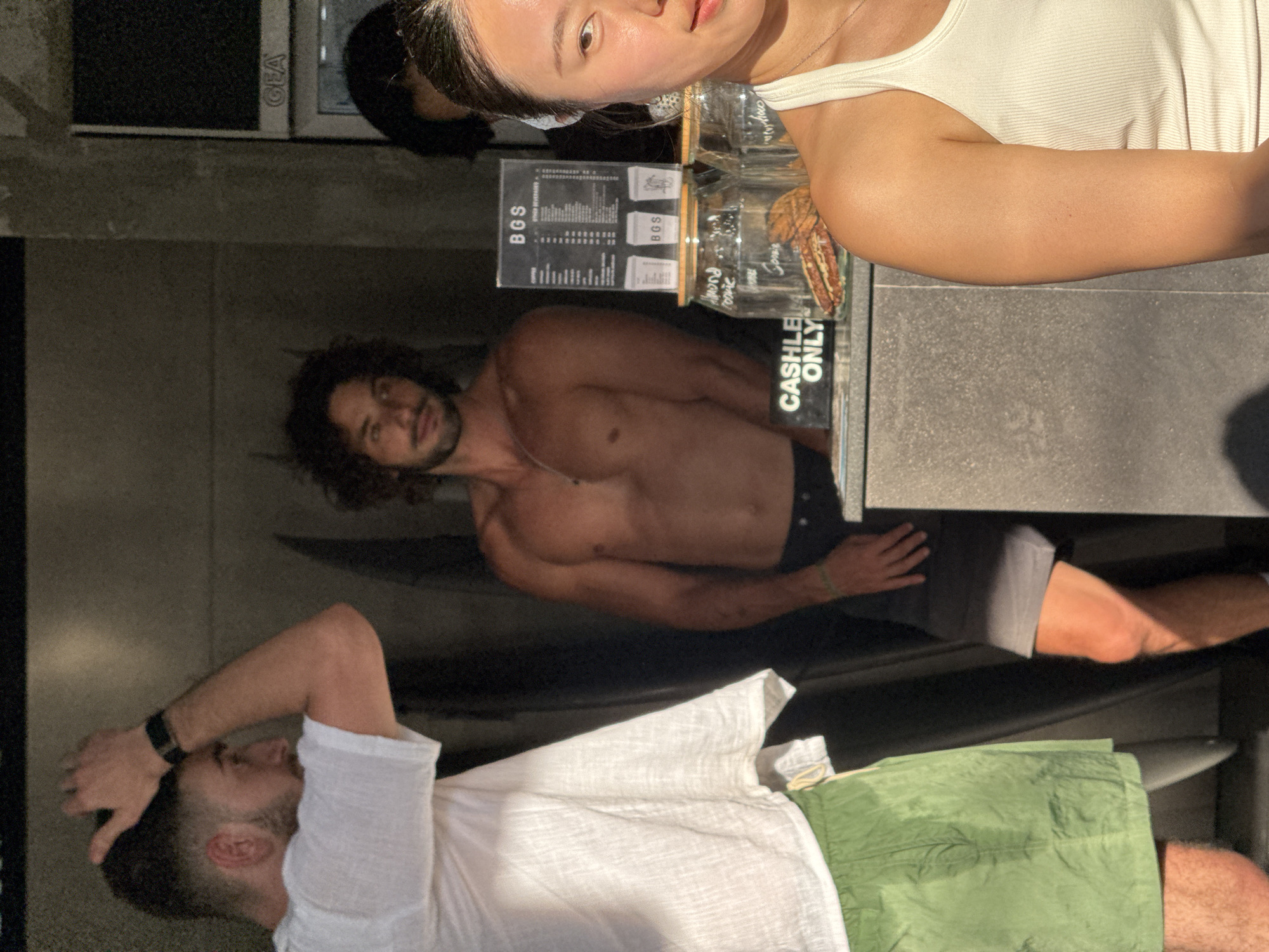The image shows three people in North Kuta, Indonesia. A woman in a white tank top stands behind a counter with a "BGS" menu and jars filled with food items. The counter reads "CASHLESS ONLY". Below the counter and to the left is a shirtless man with dark curly hair and a muscular build. To the lower left of the frame is a man wearing a white shirt and green shorts, his hand near his head. The environment appears to be a casual outdoor dining or bar area.
