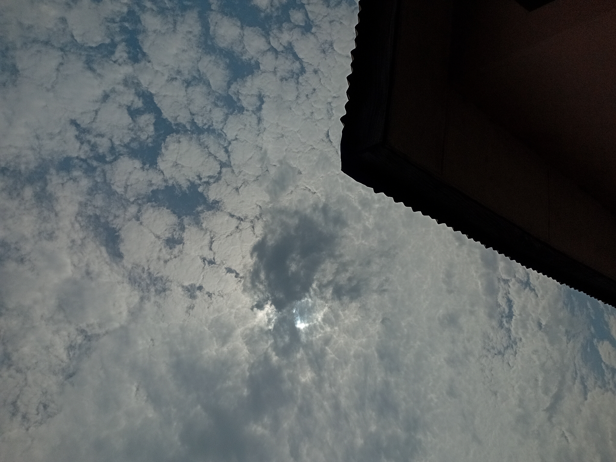The image shows a sky filled with altocumulus clouds. The sun's light pierces through a gap in the cloud cover, creating a bright spot. In the upper right corner, a portion of a dark structure, possibly a roof overhang, is visible. The location is unspecified.