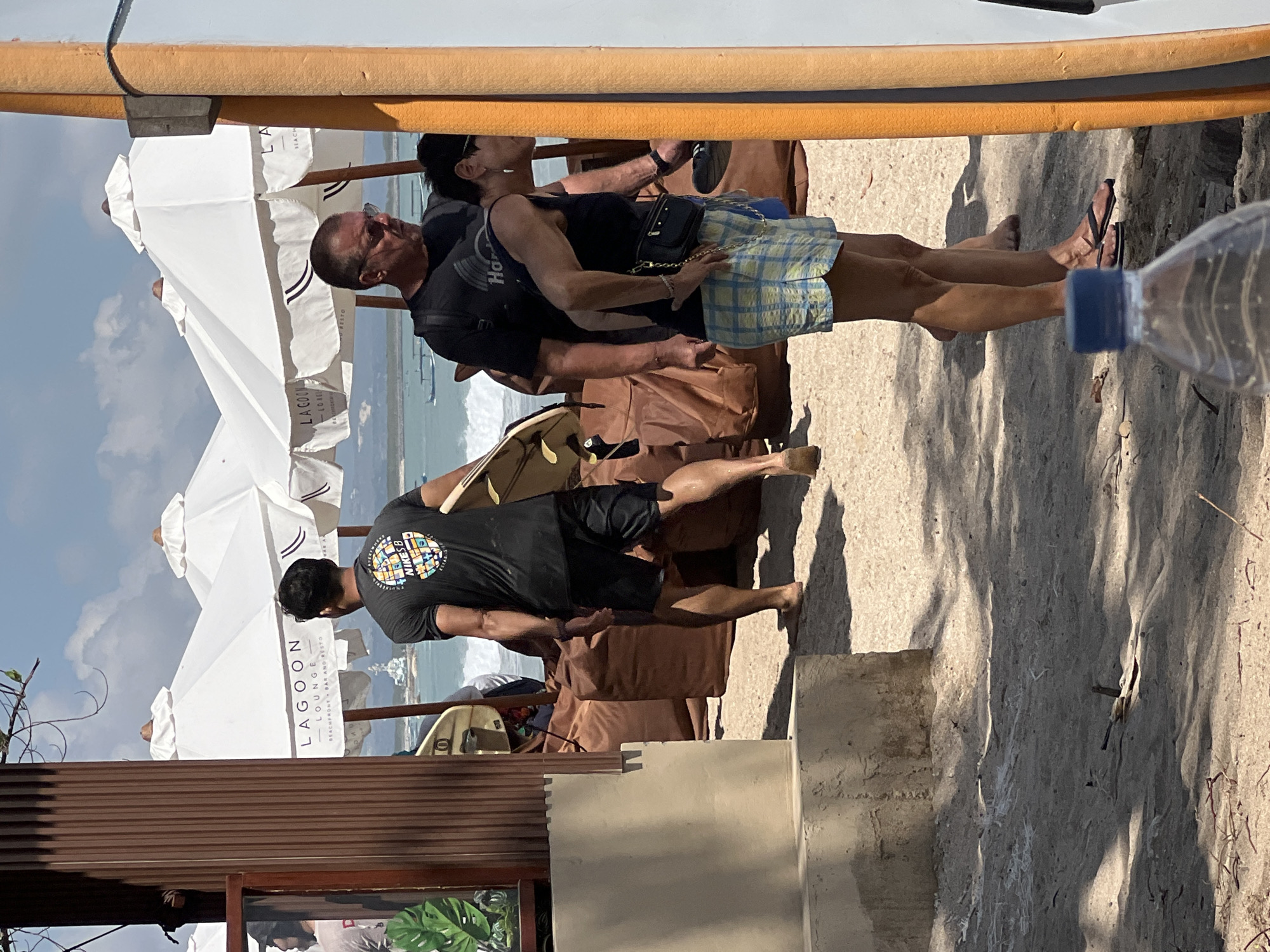 The image shows a beach scene in Kuta, Indonesia, with several people and umbrellas. In the foreground, a man in a black t-shirt and shorts walks away from the camera, carrying a surfboard. Behind him, a man with glasses and a black t-shirt stands with a woman wearing a dark tank top and plaid shorts. Both are looking towards the right side of the frame. Several white beach umbrellas are visible in the background, some with "LAGOON LOUNGE BEACHFRONT + BAR AND RESTO" printed on them. The beach is sandy, with the ocean and some boats visible in the distance. The scene appears to be set on a sunny day.