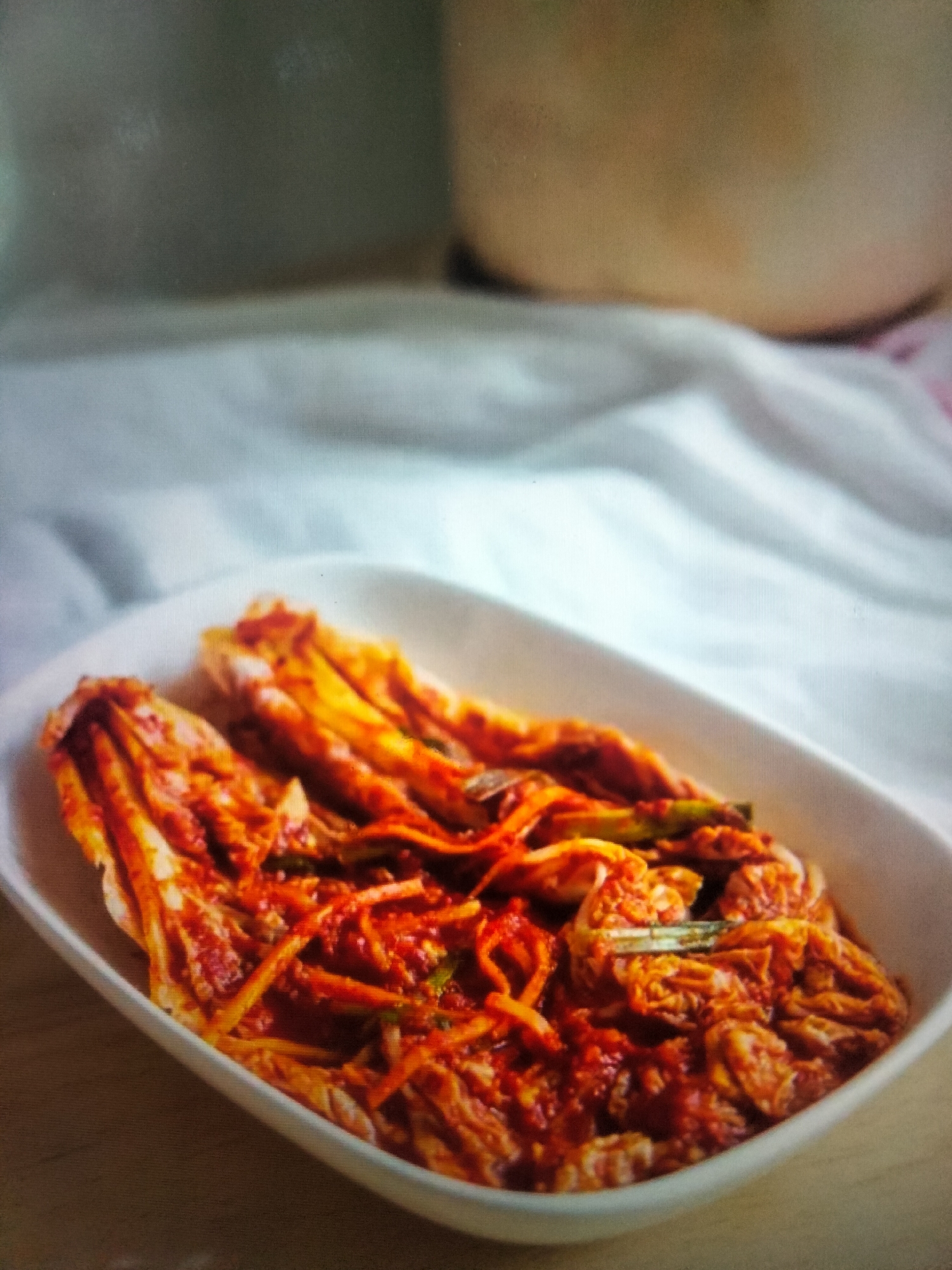 A close-up, eye-level shot captures a white, oblong bowl filled with kimchi, a traditional Korean fermented dish. The kimchi is a vibrant orange-red color, with visible napa cabbage leaves and shredded daikon radish mixed with a spicy paste. The bowl is placed on a light-colored, textured surface, likely a tablecloth, which is draped and folded in soft curves, creating a gentle backdrop. The upper portion of the image is out of focus, revealing a soft, warm-toned blur that could be a larger object or surface in the background. There is no indication of the city of Payagaboh, Indonesia, in the image.