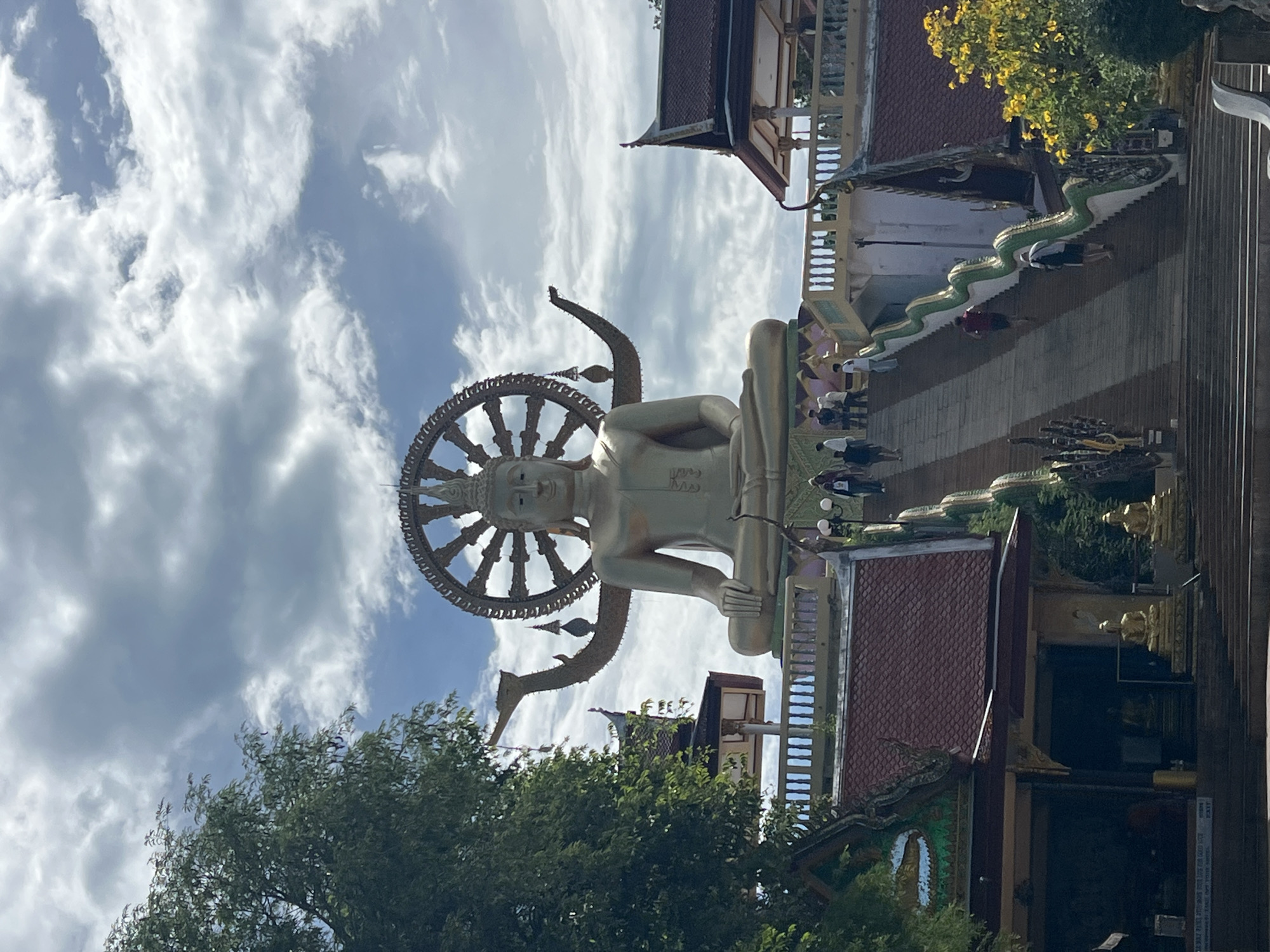 The image depicts the Big Buddha of Koh Samui, located in Baan Plai Laem, Thailand. A colossal golden statue of Buddha is seated with his legs crossed, adorned with a large, ornate wheel above his head, symbolizing the Wheel of Life. The statue is positioned on a platform, and in front of it, a wide staircase with ornate, serpentine balustrades leads downwards. Several individuals are ascending and descending the stairs. The surrounding architecture features traditional Thai temple roofs with intricate details. Lush green trees frame the left side of the image, and the sky is partly cloudy.