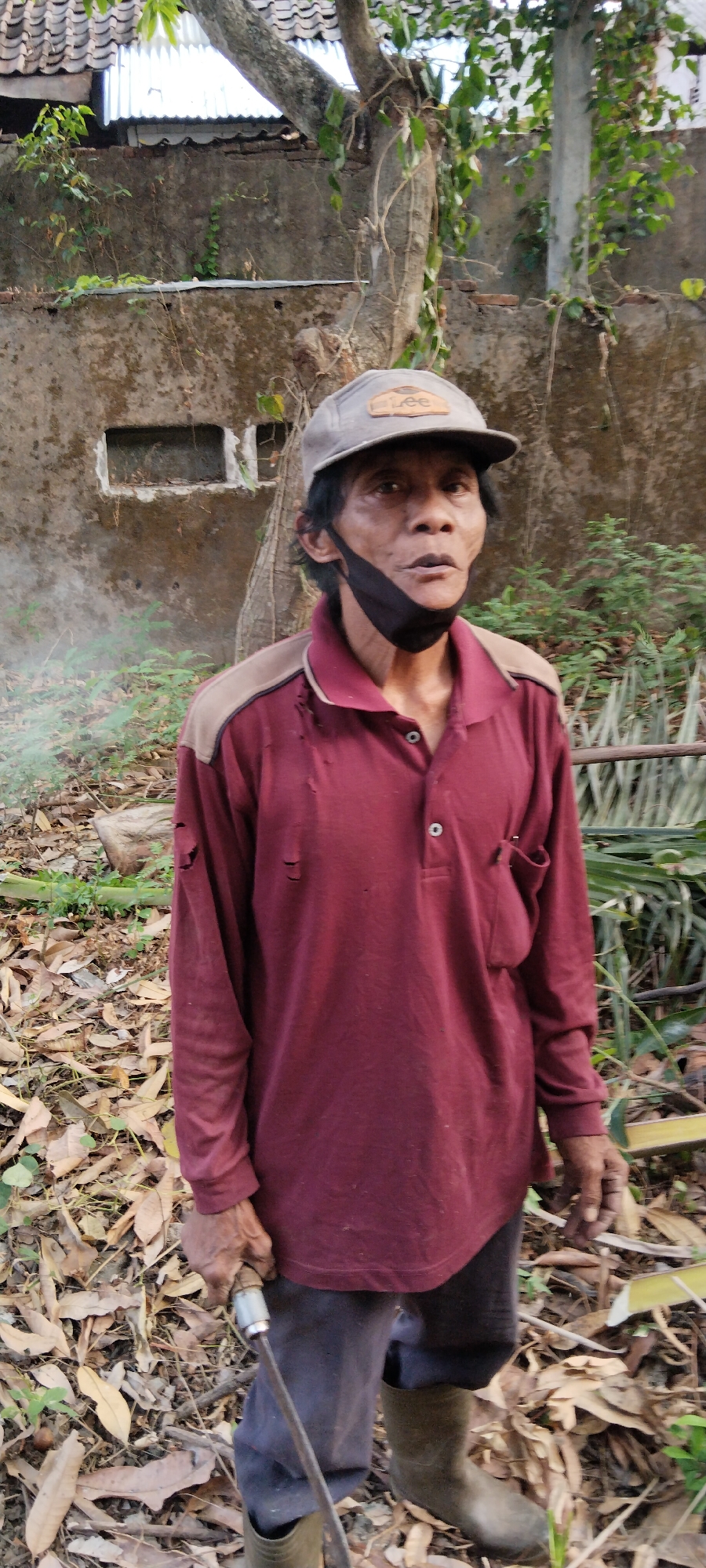 A man stands outdoors in a disheveled state, holding a tool. He is wearing a maroon long-sleeved polo shirt with tan accents on the shoulders, ripped in several places. A black mask is pulled down under his chin. He also wears a light gray baseball cap with a patch that reads "LEE." His pants are dark gray, and he has on olive-green rubber boots. The man appears to be in a rural or overgrown area, with dry leaves and vegetation on the ground. In the background, there is a rough concrete wall and a tree. To the right, there are what appear to be large palm leaves or similar vegetation. A thin plume of white smoke or mist is visible to the left of the man. The scene is consistent with the environment found in Genuk Barat, Indonesia, an area known for its agricultural and less urbanized landscape.