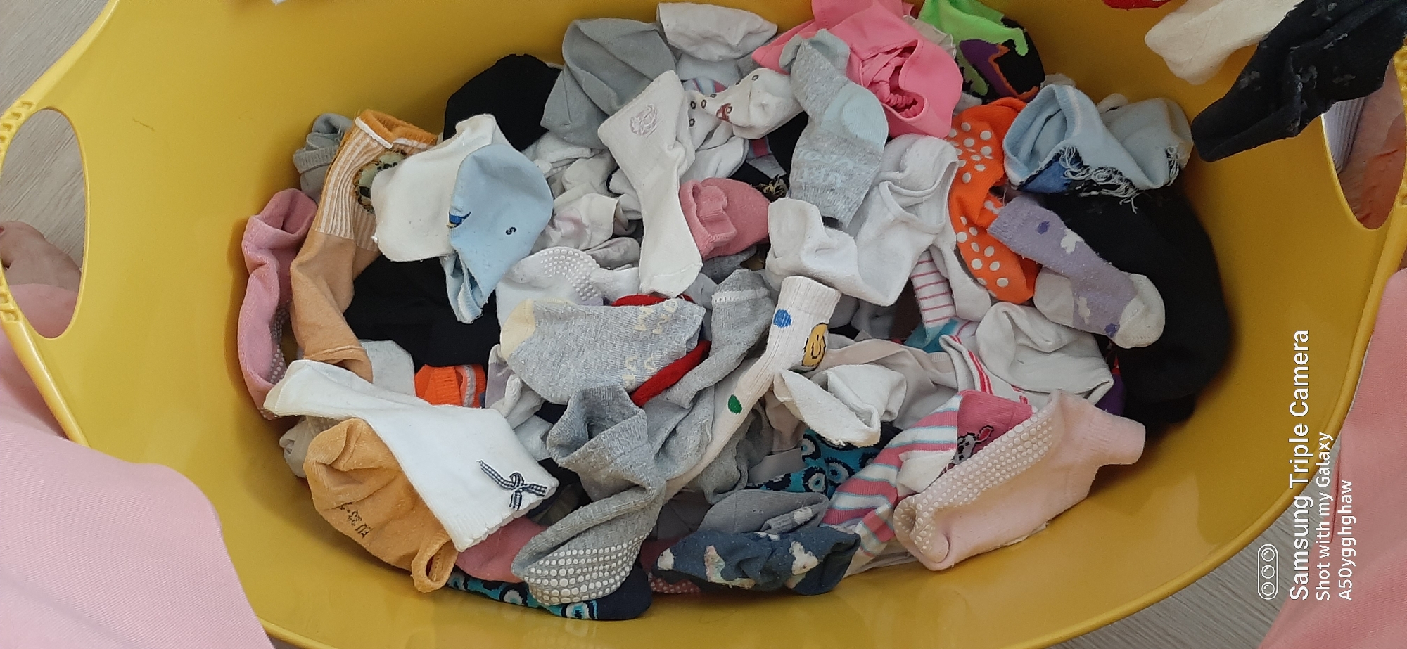 A yellow plastic laundry basket is filled with a jumble of various socks. The socks are in different colors, patterns, and sizes, suggesting they are likely a mix of adult and child socks. Some socks appear to be clean, while others may be used. The basket is placed on a light-colored floor, possibly laminate or wood. A portion of a pink fabric, possibly clothing, is visible in the upper left and lower left corners, draped over the edge of the basket. The background is indistinct, but appears to be an indoor setting. There is no indication of the location being Dubai, United Arab Emirates; this information is extraneous to the visual content of the image.