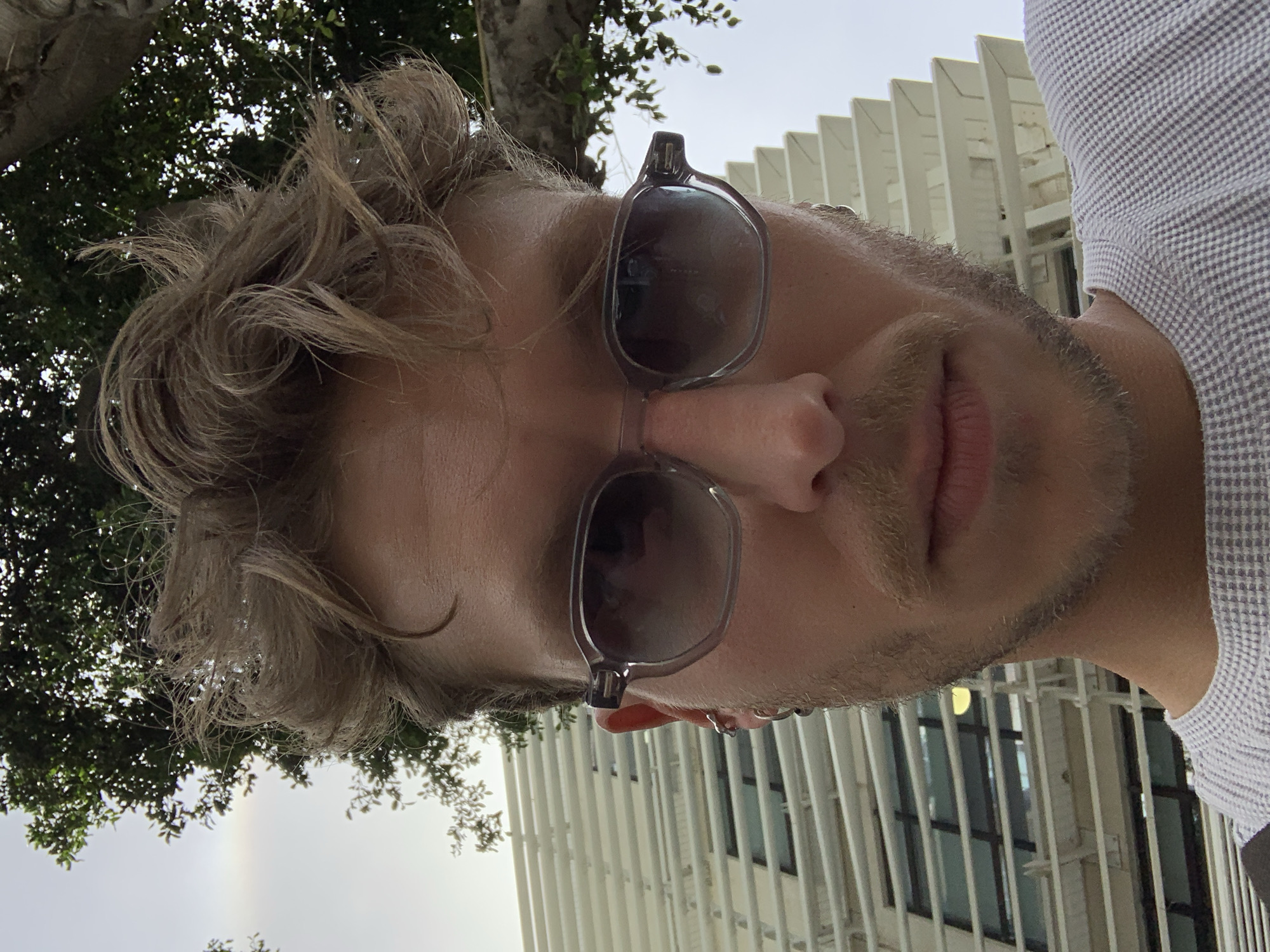 A close-up, selfie-style photograph shows a man with fair skin, light brown hair in a disheveled style, and a beard with a mustache. He is wearing sunglasses with gray frames and dark lenses, which are obscuring his eyes. A light-colored, checkered collared shirt is visible on his right side.
In the background, on the left, are tree branches with green leaves against a bright, overcast sky, possibly exhibiting a faint halo effect. On the right, a building is visible with a distinctive architectural design featuring vertical, white fins or beams, possibly part of a modern structure.
Given the architectural style and general atmosphere, the location is possibly within Tel-Aviv, Israel.