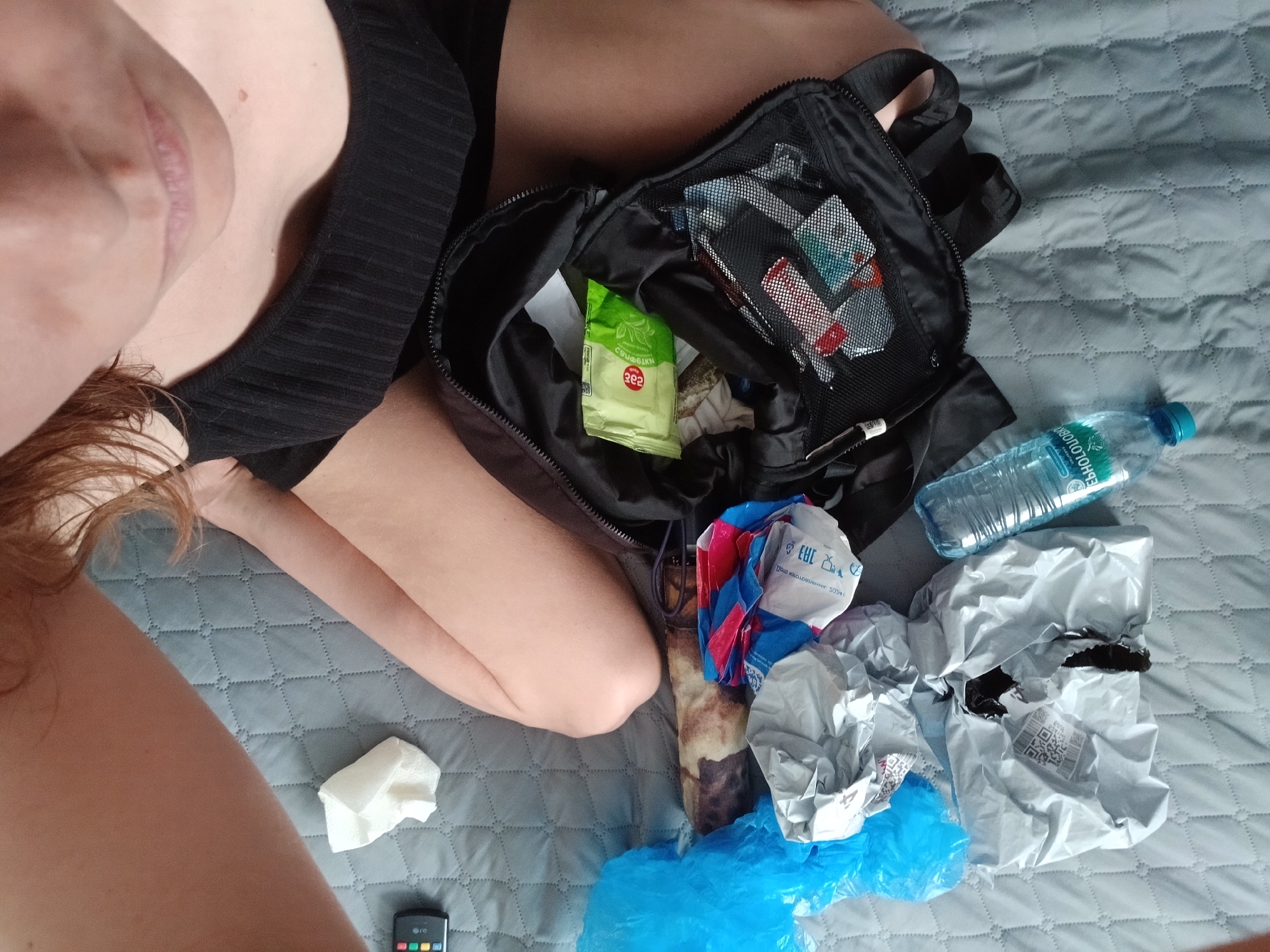 The image depicts a high-angle, close-up view of a person's upper body and a collection of items on a quilted gray surface, likely a bedspread. The person, with fair skin and reddish-brown hair, is wearing a black ribbed tank top. Their face is partially visible from a low angle, showing their mouth and chin.
Scattered around the person are various objects. To the left, a crumpled white tissue and a black remote control with colored buttons are visible. A bright blue plastic bag is also present, next to a long, brown, and beige patterned object, possibly a wrapped item or a cylindrical container. Several other crumpled plastic bags in shades of blue, red, and white are clustered together, some with visible text and logos. One white crumpled bag has text that appears to be in Cyrillic, including "ЕРС" and "3054".
In the center and right of the image, a black backpack is open, revealing its contents. Inside, there is a green package of wet wipes with Cyrillic text, including "САЛФЕТКИ" and "362". A mesh compartment within the backpack contains several small, rectangular packages with red and blue detailing, possibly medications or other personal care items. A clear plastic water bottle with a blue cap and green text is lying on its side to the right of the backpack. The text on the bottle is also in Cyrillic, including "БНОЛОГ", "МИНЕРАЛЬНАЯ", and "ВОДА".
The overall scene suggests a disorganized or recently unpacked collection of personal belongings and purchases. The presence of Cyrillic text on some items indicates a possible location in Russia. Based on the Cyrillic text and common packaging found in Russia, it is plausible that this scene is located in the city of Yekaterinburg, Russia, although this cannot be definitively confirmed solely from the visual information provided.