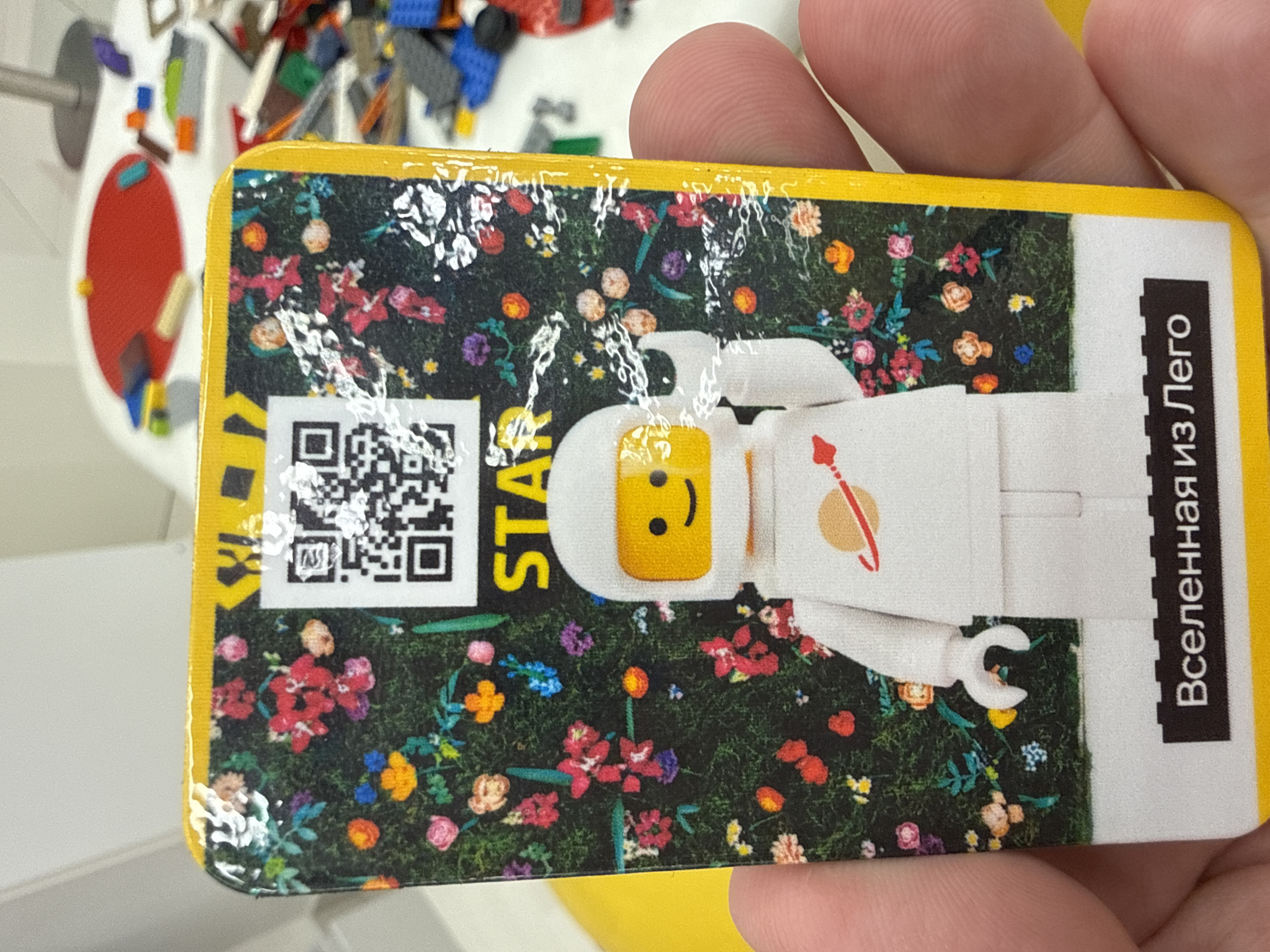 The image shows a hand holding a rectangular card. The card features a colorful floral background with a QR code and the word "STAR" printed on it. Below the QR code is an illustration of a Lego astronaut with a smiley face and a white suit with a planet emblem. At the bottom of the card, in a black rectangular box, is the Russian text "Вселенная из Лего" which translates to "Universe from Lego". The background of the image is slightly blurred but appears to be an indoor setting with some Lego building blocks scattered on a table in the distance. The card is likely a collectible item or part of a game. There is no specific information in the image that indicates the location in the city of Khimki, Russia.
