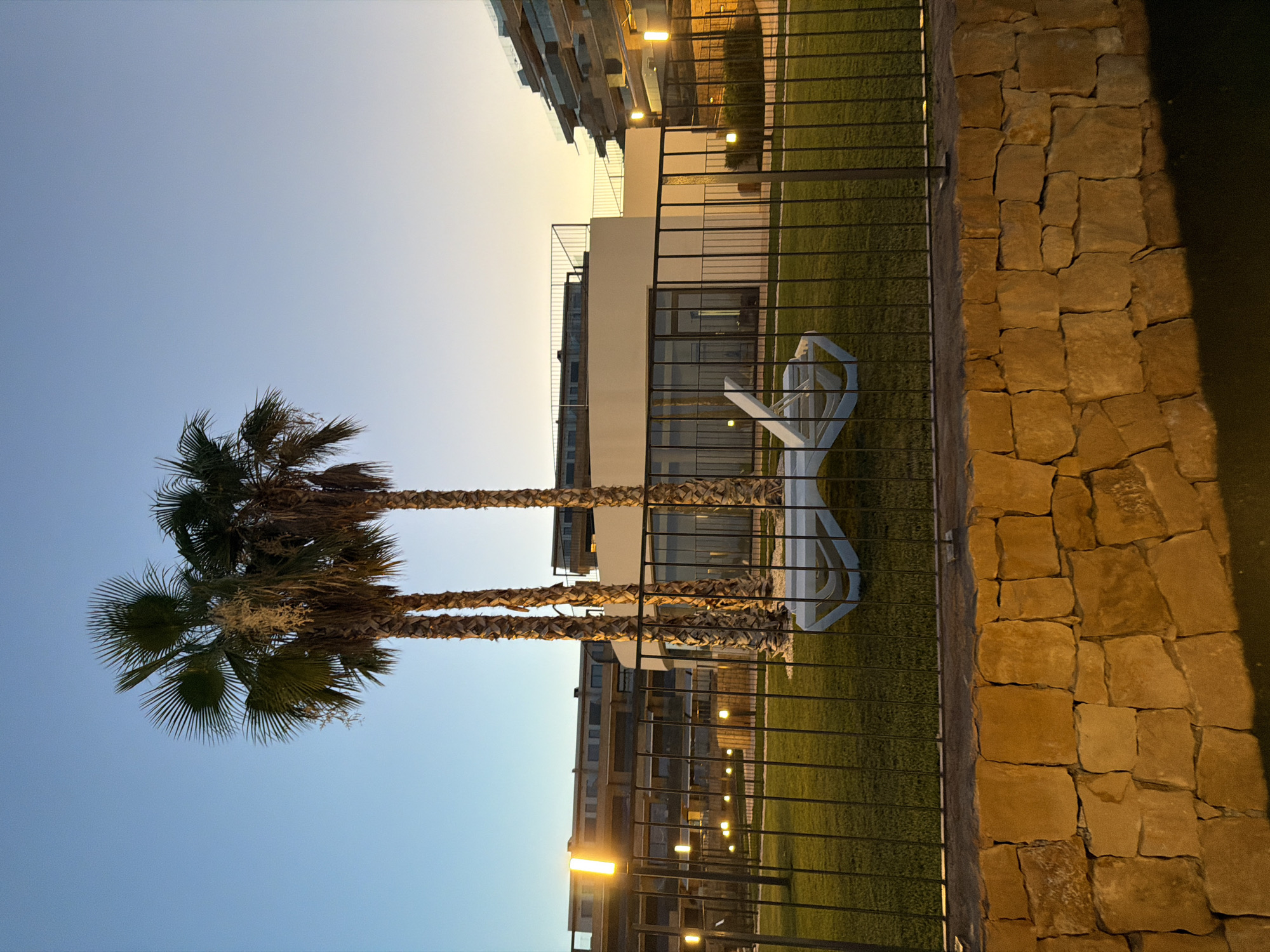 The image depicts a twilight scene in Finestrat, Spain, featuring a resort or residential complex. In the foreground, a low, rough-hewn stone wall separates the viewer from a manicured lawn enclosed by a black metal fence. Three palm trees stand prominently on the lawn, their fronds silhouetted against a gradient sky transitioning from clear blue at the top to a warm yellow-orange near the horizon. A white, modern lounge chair is visible on the grass. Beyond the fence and palm trees, several buildings are visible. To the left, a long, multi-story building with many windows can be seen, with orange outdoor lighting illuminating parts of the facade. In the center, a curved, white building with large glass windows forms a central structure. To the right, another multi-story building with a unique, stepped architectural design, featuring balconies with glass railings, rises into the sky, also illuminated by warm lighting. The overall impression is one of a modern, upscale leisure or residential environment at dusk.