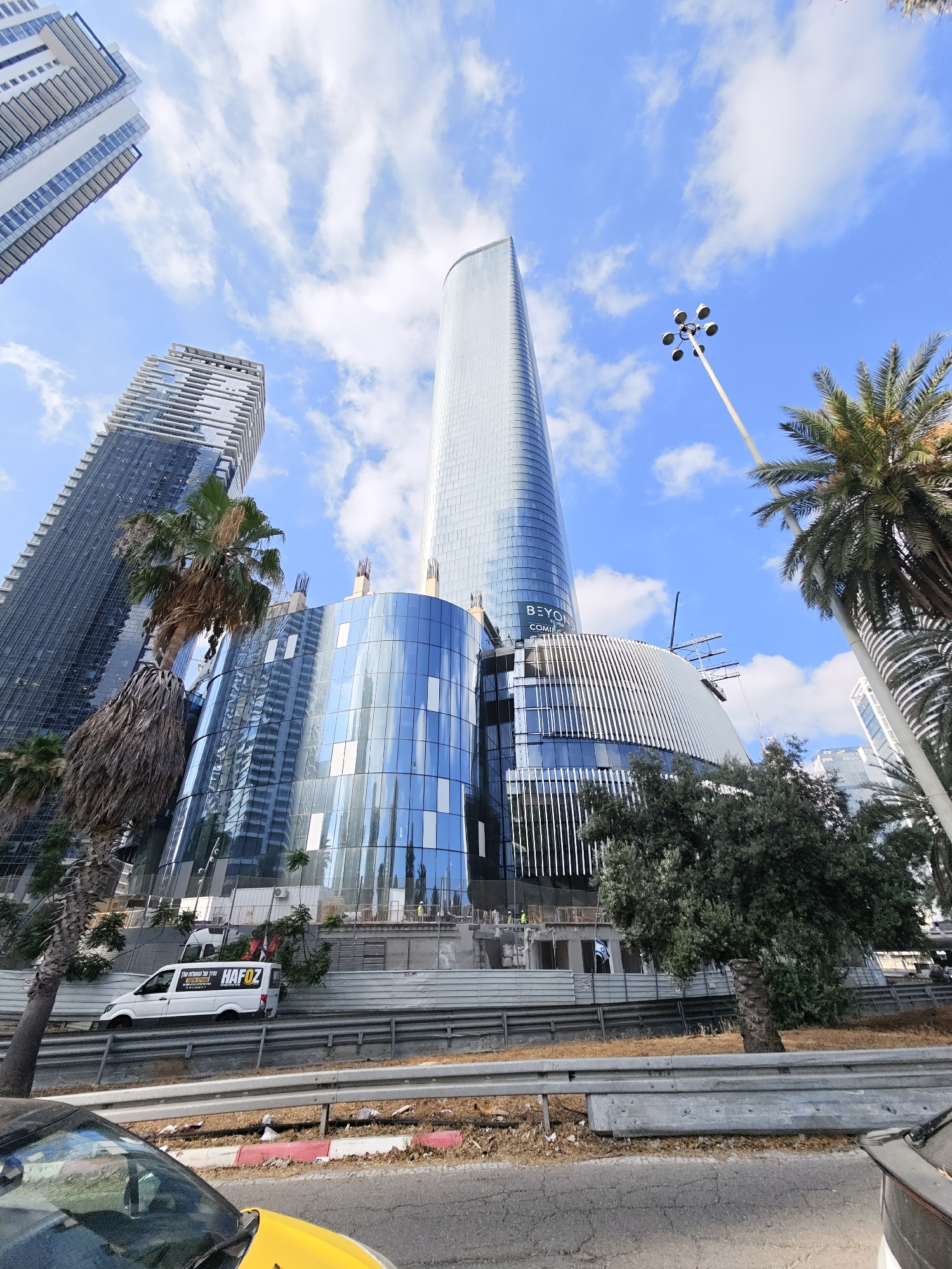 A low-angle, wide-shot photograph taken on a bright, partly cloudy day in Givatayim, Israel, captures the modern architecture of multiple high-rise buildings, including a distinctive curved glass structure that appears to be under construction. In the foreground, a white van with the logo "HAFOZ" is visible parked behind a metal barrier. The van has text in Hebrew and English on its side, which reads "HAFOZ IMPORT AND EXPORT." The surrounding environment includes palm trees, vegetation, and a street with visible road markings and debris. The architectural style of the buildings suggests a contemporary urban setting, with sleek lines and extensive use of glass. A tall street lamp with multiple bulbs stands to the right of the frame, and scaffolding is present on the construction site.