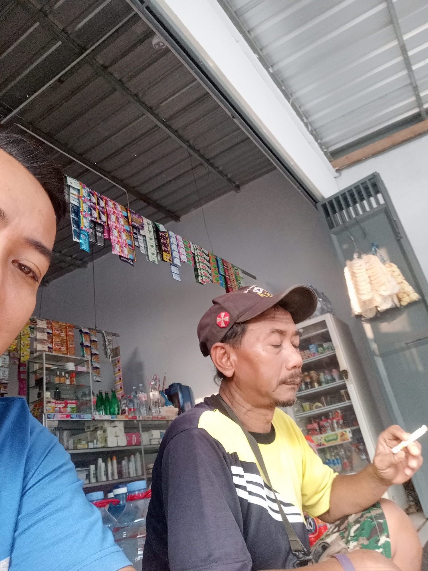 In Simo, Indonesia, a man wearing a brown baseball cap and a yellow and dark blue shirt is seen from the chest up, holding a lit cigarette in his right hand. He has a mustache and a goatee. He appears to be seated in front of a shelf filled with various products, possibly in a small shop or convenience store. To his left, another person is partially visible, wearing a blue shirt. The background shows a ceiling made of corrugated metal and a wall with shelves stocked with items. There are also some hanging items in the background, resembling snack packaging and what appear to be crackers. The lighting suggests it is daytime.
