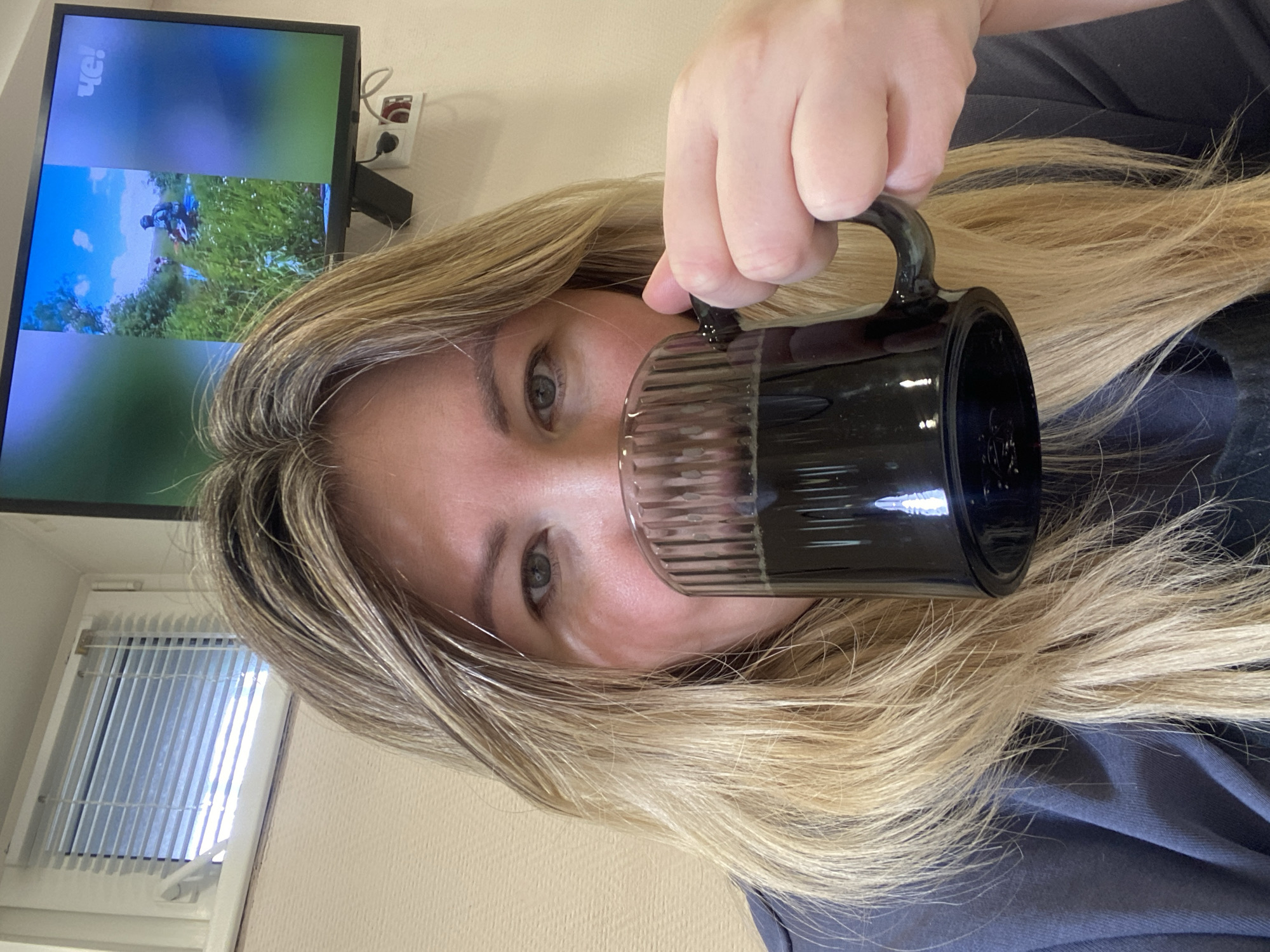 The image, rotated 90 degrees clockwise, features a woman indoors, holding a dark, fluted glass mug to her mouth. She has long, light blonde hair that partially covers her face and shoulder. Her eyes, with a neutral expression, look towards the viewer. She is wearing a dark-colored top.
In the background, to the left of the woman's head, a television screen displays an outdoor scene with green grass, a blue sky, and a person in what appears to be outdoor attire. The logo "ЧЕ!" (Che!), indicating a Russian television channel, is visible in the upper left corner of the screen. A power outlet with a plugged-in cord is mounted on the wall behind the TV. Below the television, a window fitted with white vertical blinds is visible, confirming an indoor setting. The scene suggests a casual and relaxed moment.