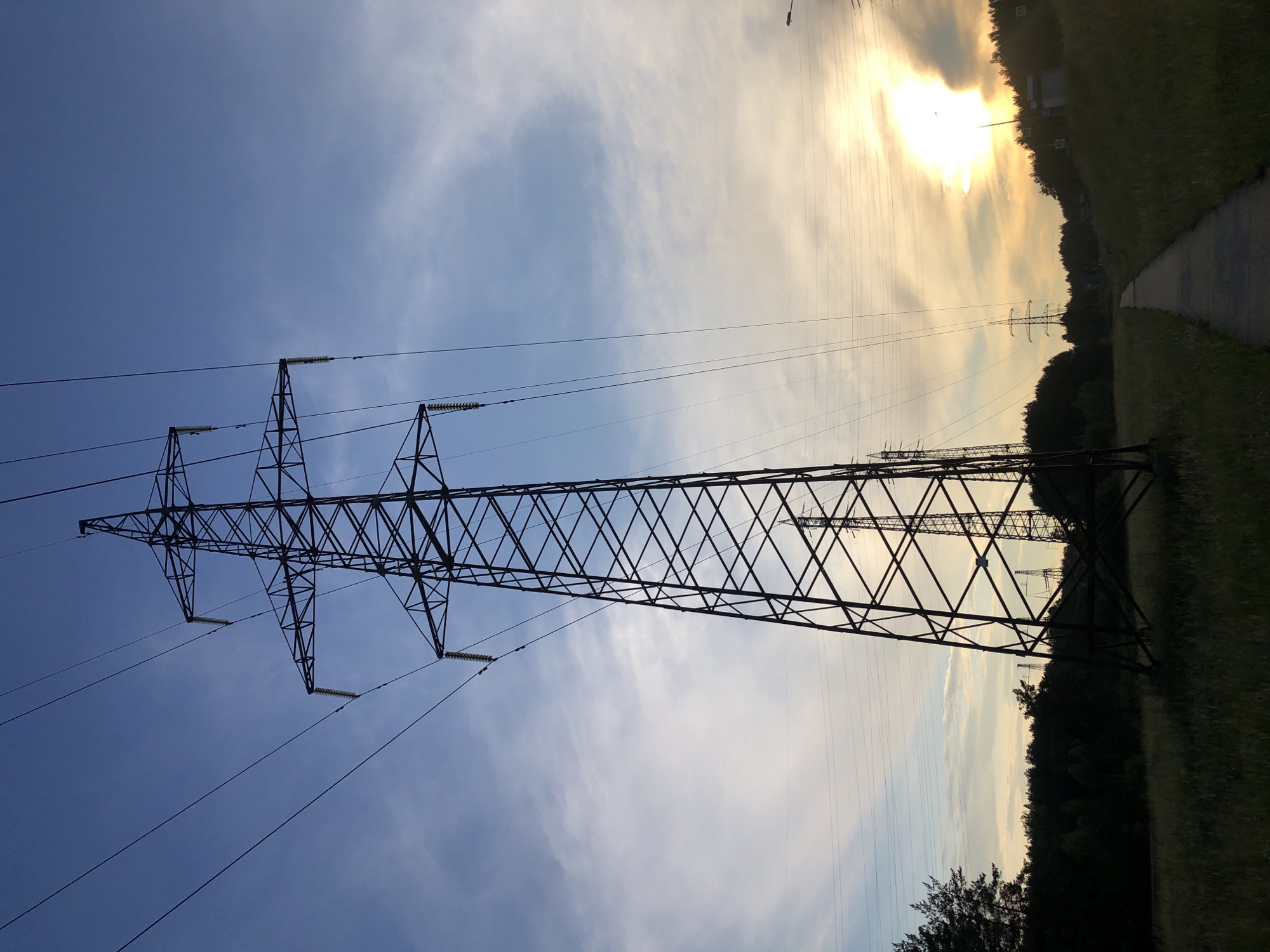This is a landscape photograph featuring a series of electricity pylons under a sky with mixed clouds. The sun is visible in the upper right portion of the image, casting a warm glow and creating a sunset or sunrise effect. The pylons are tall, lattice-like metal structures supporting thick power lines that stretch across the frame. The foreground is dominated by a large pylon, viewed from below, emphasizing its immense size and geometric structure against the sky.
In the background, more pylons can be seen receding into the distance. To the right, a grassy slope leads to a path, and beyond that, a line of trees forms a dark silhouette against the brighter sky. The overall scene suggests an open, possibly rural or semi-rural, environment. The time of day is clearly late afternoon or early evening due to the low angle of the sun and the quality of the light. There is no visible text in the image. The mood is somewhat dramatic due to the stark silhouettes of the pylons against the illuminated sky.