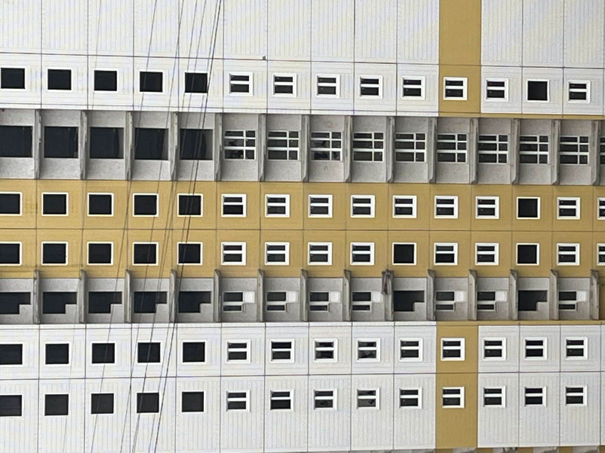 The image depicts a close-up of a building facade, likely an apartment complex, in an unidentified urban location. The building is characterized by a repetitive pattern of rectangular windows arranged in horizontal rows. The color scheme alternates between white, gray, and yellow, defining the different levels or sections of the building. The windows vary in style, some being simple square openings, others featuring balconies or recessed structures, and yet others divided into smaller panes. Several thin wires or cables run diagonally across the facade, adding a layer of complexity to the composition. The overall aesthetic is minimalist and utilitarian, suggesting a functional architectural design. There are no discernible people or signs of street-level activity.