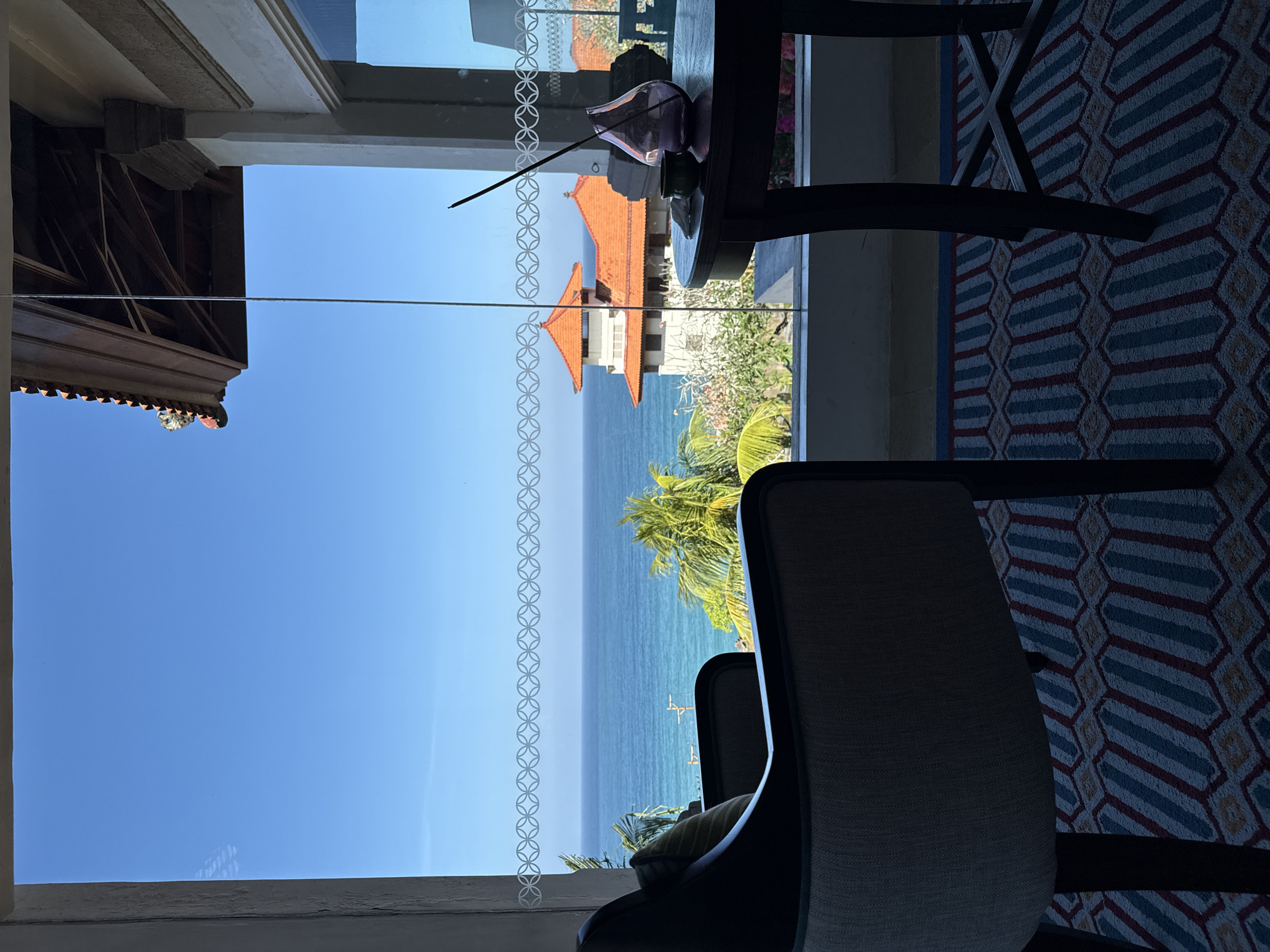 The image depicts an indoor scene looking out through a large window at a tropical ocean view in Kuta Selatan, Indonesia. In the foreground, a dark upholstered armchair with a patterned cushion is positioned on a rug with a geometric design in blue, red, and beige. To the right, a dark wooden round table holds a purple glass vase with an incense stick and a small green dish. Through the window, a clear blue sky meets a turquoise ocean. In the middle ground, lush green palm trees and other tropical foliage are visible. Beyond the vegetation, a building with traditional orange-tiled roofs and white walls, characteristic of Balinese architecture, stands prominently. The water exhibits calm conditions with gentle ripples. The sun appears to be shining brightly, casting shadows within the room. The scene suggests a serene and luxurious setting overlooking the ocean.