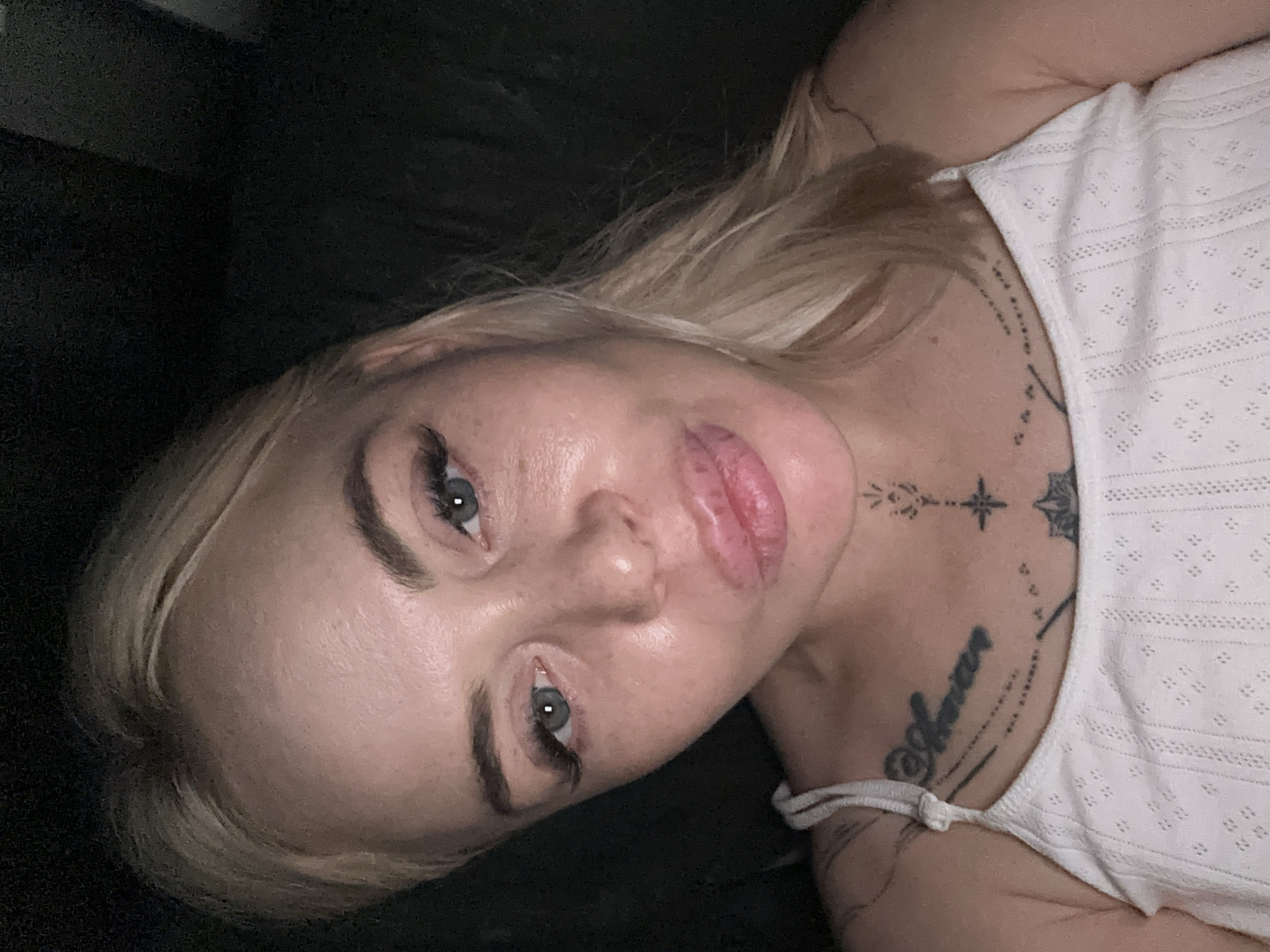 The image presents a close-up, frontal portrait of a female individual with fair skin and blonde hair, which is parted slightly off-center and falls over her shoulders. Her eyes, appearing blue or grey, are directed towards the camera, and she has visible eyelashes, possibly enhanced. Her eyebrows are defined. Her lips are full and slightly pursed or puckered. The skin on her face exhibits natural texture, including visible pores and minor variations. She is wearing a white, textured sleeveless top with thin straps.
Multiple tattoos are visible on her upper chest and lower neck. On the viewer's left side of her chest, a script tattoo is present, with some letters appearing to form part of a name or word. Centered on her upper sternum/lower neck is a vertical tattoo design incorporating geometric elements, a star or cross-like shape, and a floral or mandala-like motif below it. Faint tattoo lines are also visible extending from the left shoulder onto the upper arm.
The background is uniformly dark and devoid of specific features, suggesting a low-light indoor setting or a plain dark surface. The lighting appears to be direct and frontal, casting minimal shadows and highlighting facial features. The location is an unknown setting within the city of Unknown, Unknown.