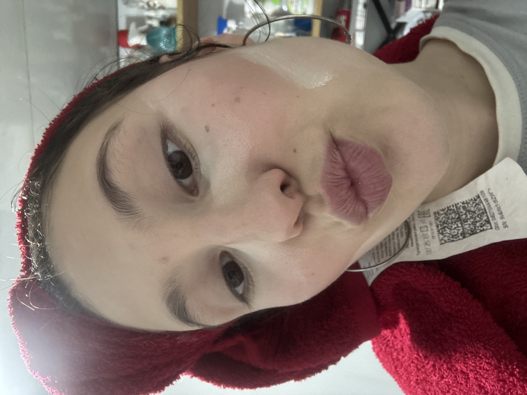 A close-up, slightly tilted portrait captures a woman with fair skin, brown eyes, and light-pink lipstick. A white, creamy substance is applied to her right cheek, possibly makeup or skincare. She is wearing a large silver hoop earring and a red, textured towel wraps around her head. A clothing tag with a QR code and text in a foreign language (likely Russian) is visible on her chest. The background is blurry, showing a bathroom shelf with various personal care items. There is no indication about being in the city of Фоки, Russia.