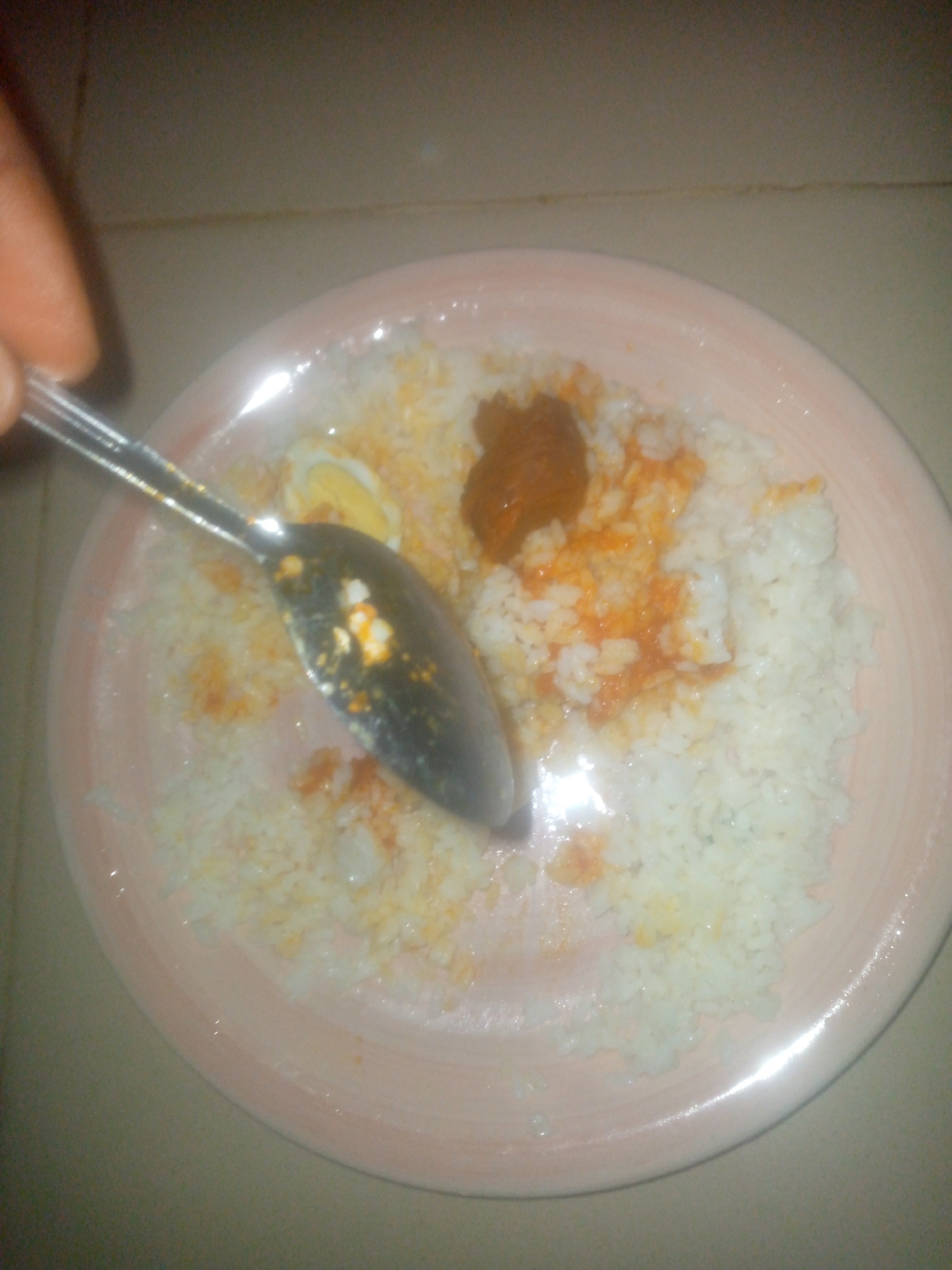 A high-angle, close-up shot reveals a partially consumed plate of food. The meal consists primarily of white rice, accompanied by a sliced hard-boiled egg and a reddish-brown sauce. A metal spoon is present, seemingly mid-action in mixing or scooping the food. The plate is a light pink color with a subtle rim design. The surface underneath appears to be a light-colored tile or countertop. The location is specified as Unknown, Unknown.