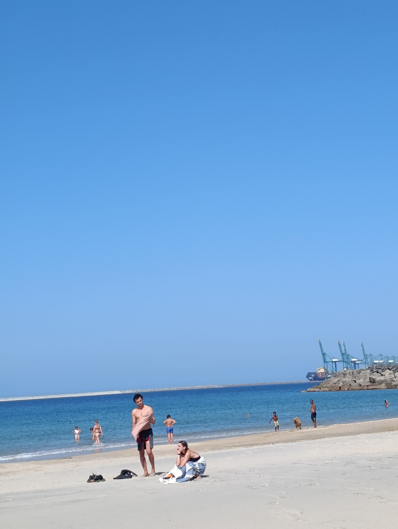 The image depicts a sunny day at the beach in Sines, Portugal, with a clear blue sky and calm ocean waters. In the foreground, a man is standing on the sand, holding a towel. Nearby, a woman is sitting on the sand, leaning back on her arms. Other beachgoers are visible in the shallow water, including children and adults. In the background, a long breakwater stretches across the ocean, and a large cargo ship and several cranes are visible at the port of Sines, with a rocky breakwater also present. The sand is light in color, and the water is a vibrant blue. Several items of clothing and accessories are scattered on the sand in the foreground.